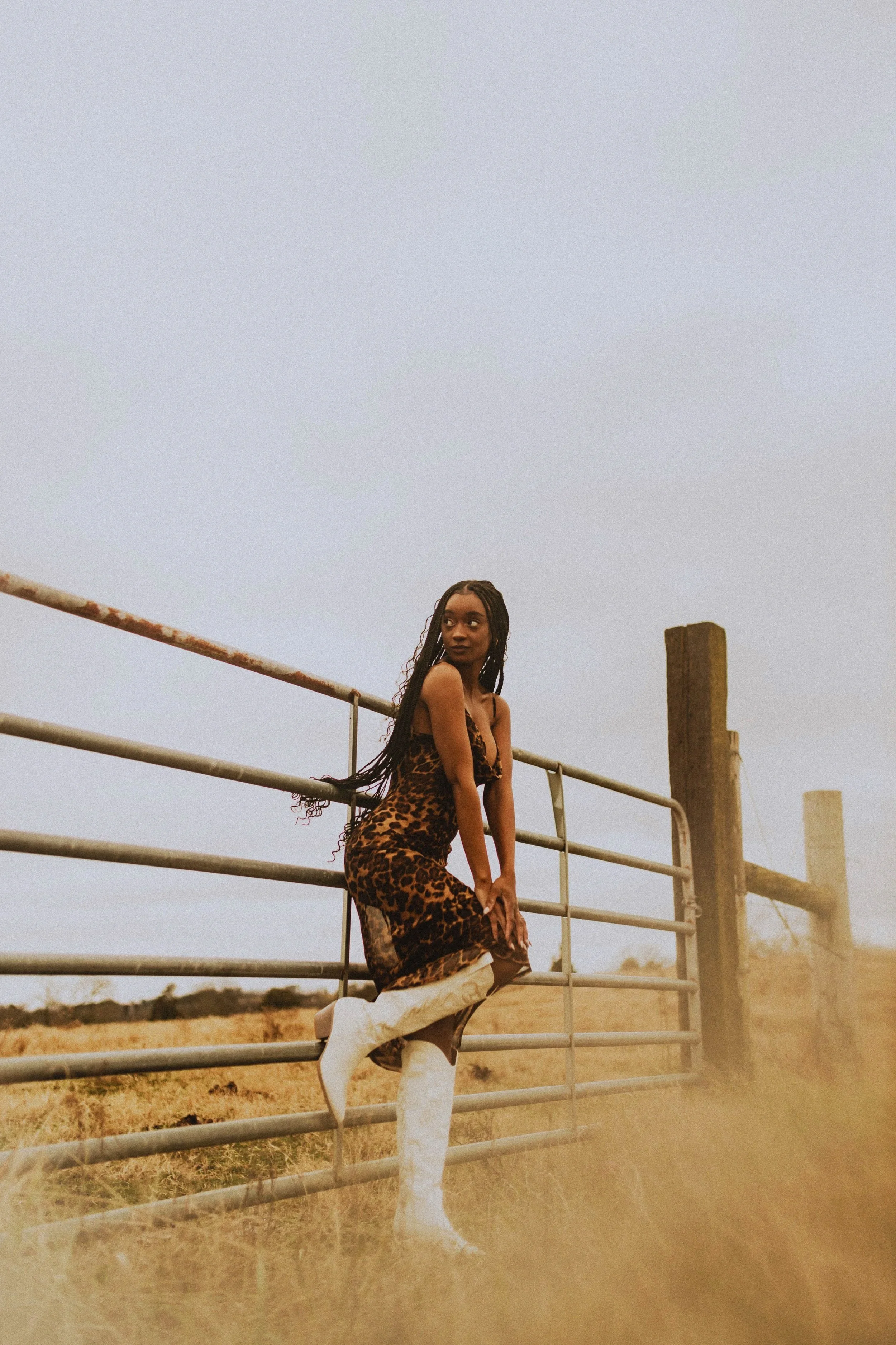A woman dressed in a leopard print dress and white knee-high boots leaning on a metal gate in a rural area with grass and wooden posts, under a cloudy sky.