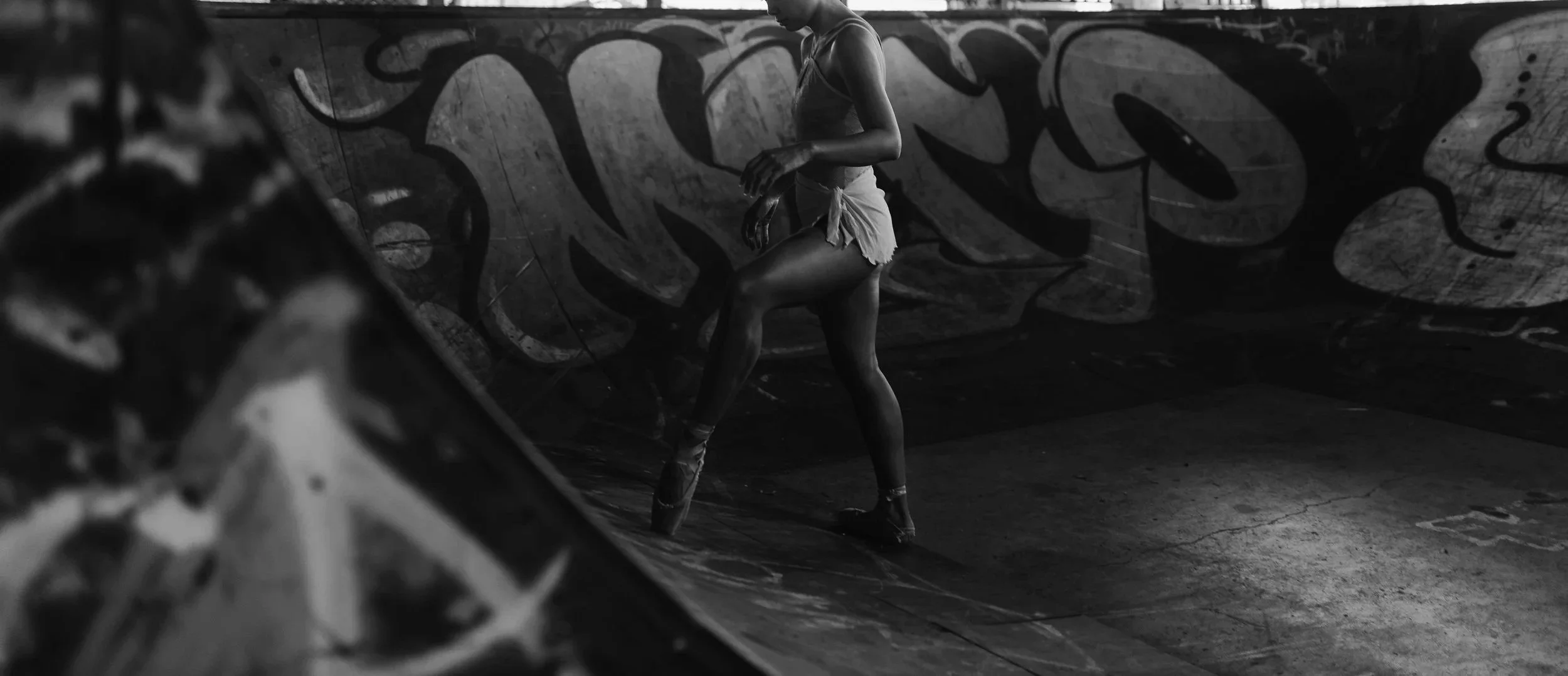Black and white photo of a ballet dancer practicing on a graffiti-covered skate ramp.