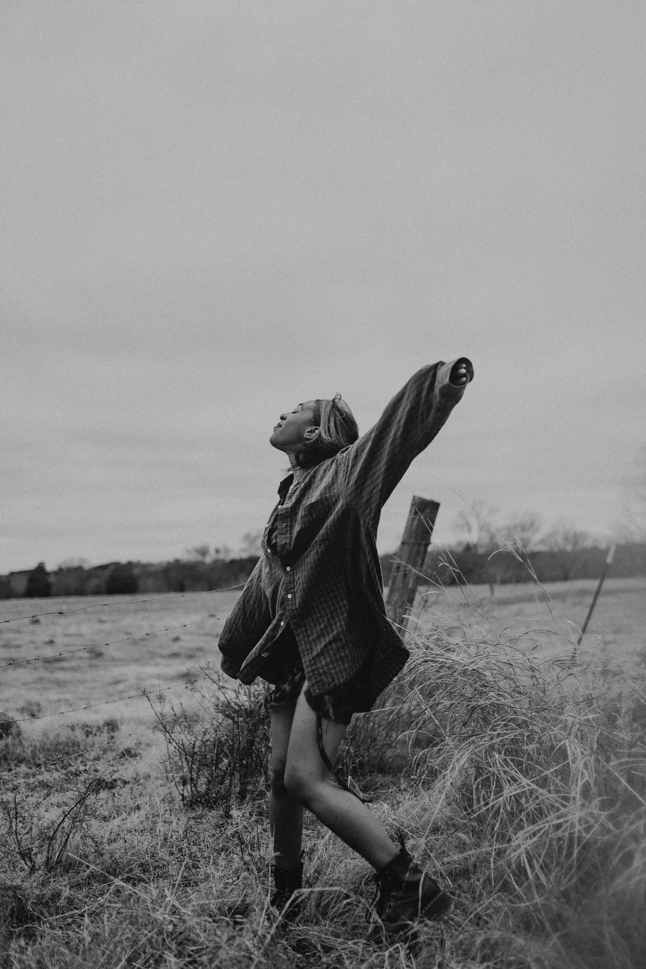 A woman stands outdoors in a grassy field with her arms outstretched, eyes closed, and head tilted back, wearing a checkered jacket and shorts, in black and white photo.