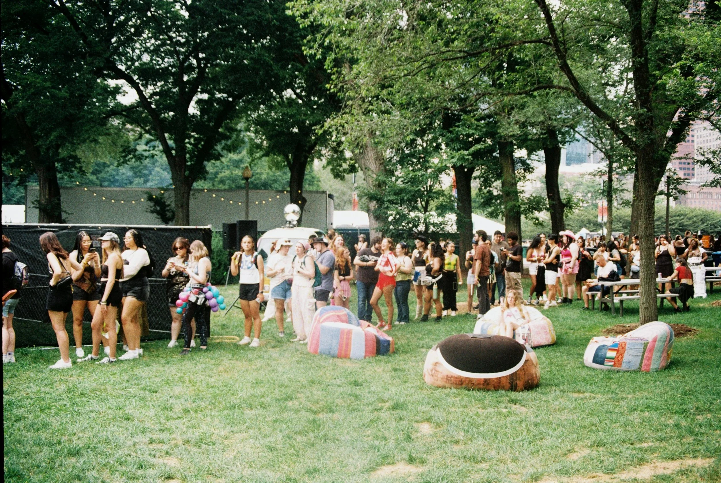 People standing and sitting in a park during Lollapalooza, with trees and city buildings in the background, at an outdoor festival By Chicago Based Photographer Taylor Tolbert | Ttolbphotog