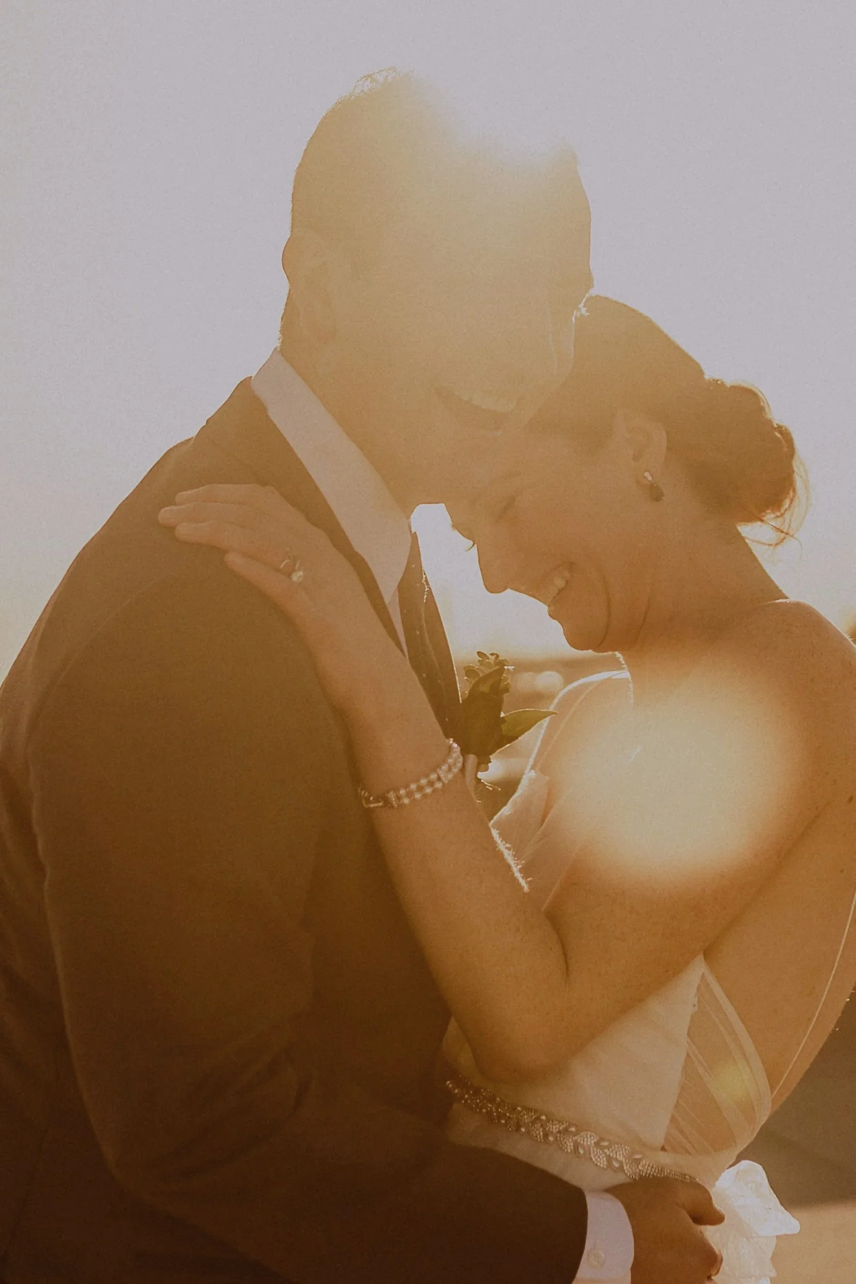 Bride and groom embracing, dressed in formal wedding attire, with sun flare creating a warm glow.