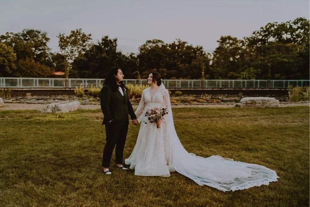 Couple in wedding attire holding hands on grass field with trees in background.