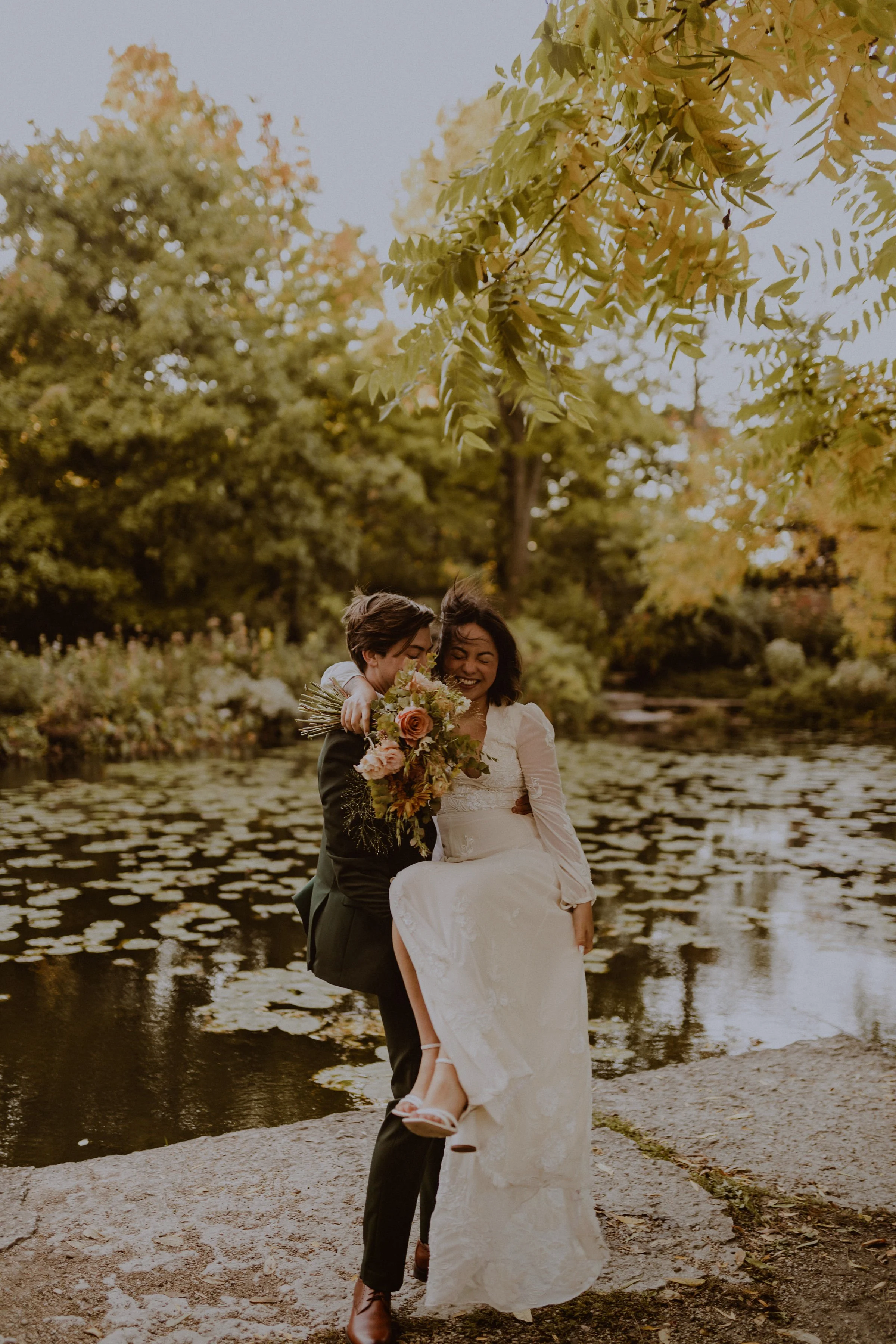 A couple celebrating in a natural outdoor setting by a pond, with one person in a wedding dress being lifted by their partner in a suit, both smiling and holding a bouquet of flowers. Image taken at the Alfred Caldwell Lilly Pool in Lincoln Park Chic