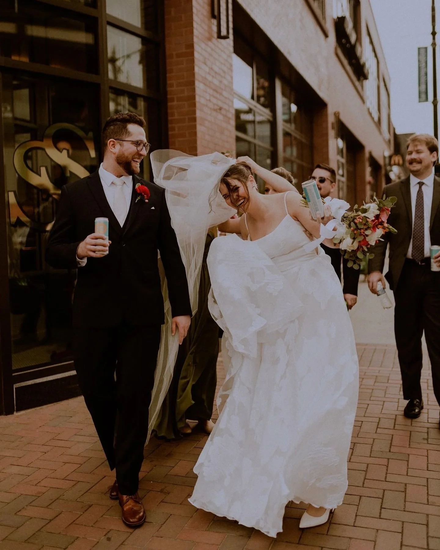 A bride and groom are surrounded by friends on a city sidewalk, celebrating their wedding as they smile and laugh. The bride is in a white wedding dress and veil, while the groom is in a black suit with a red boutonniere. Friends are holding drinks a
