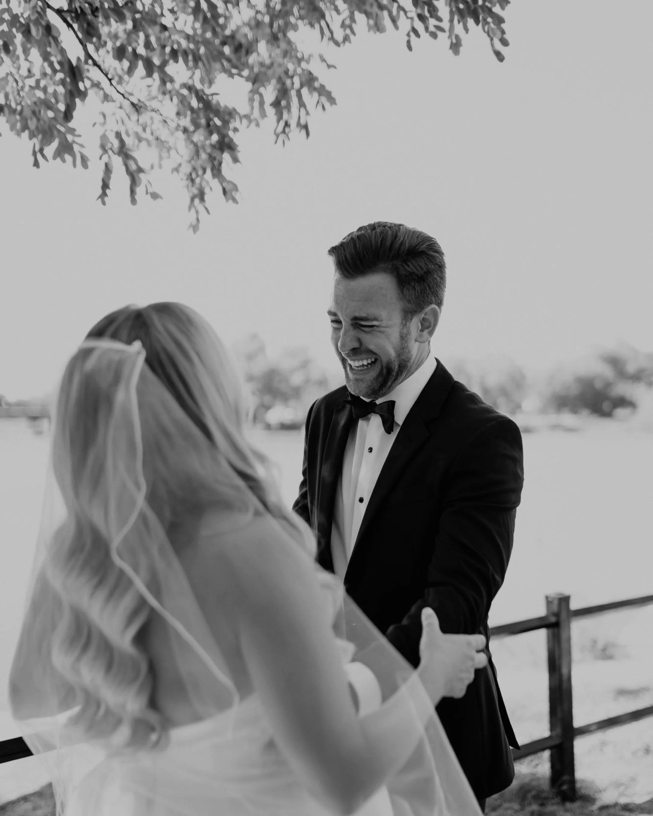 Black and white photo of a bride and groom smiling at each other outdoors, with the bride wearing a veil and the groom in a tuxedo.