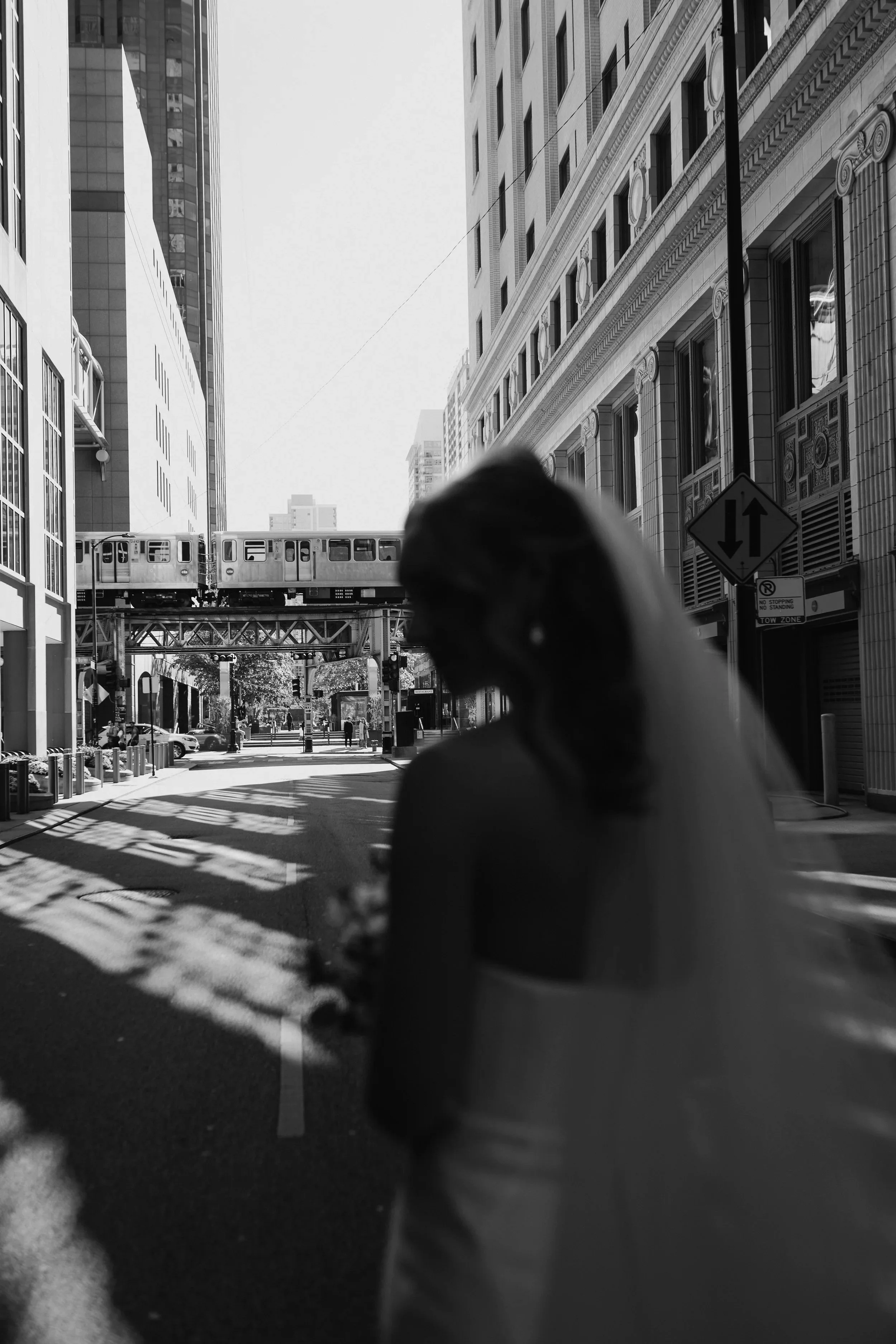Black and white photo of a silhouette bride in a city street with an elevated train passing overhead and tall buildings flanking the scene.