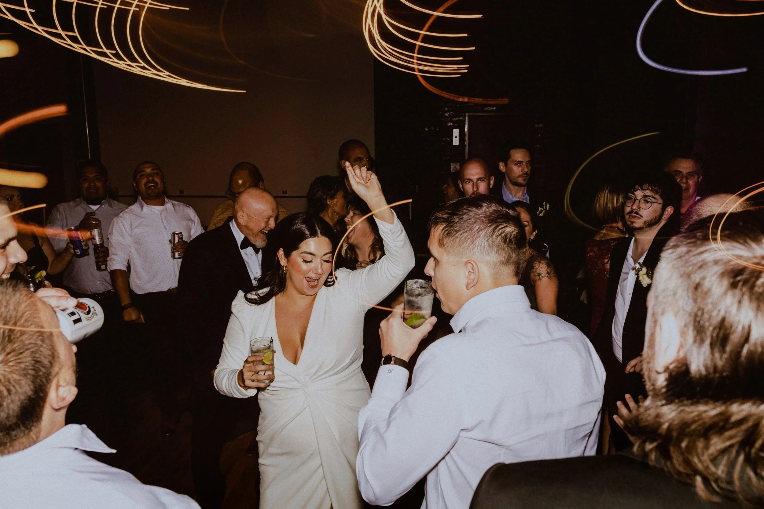 A woman in a white dress dancing and holding a drink at a lively party or celebration with many people surrounding her, some holding drinks and some dancing, in a dimly lit room with swirling light effects.
