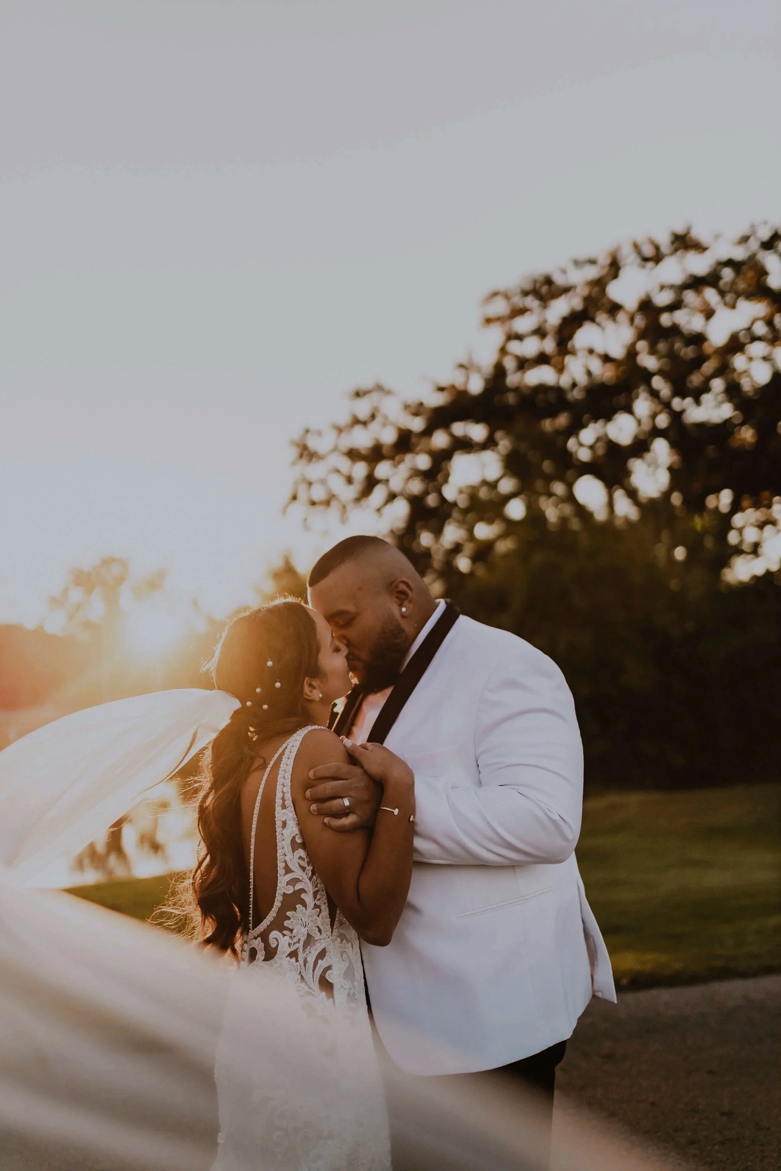 A bride and groom kissing outdoors during sunset with the bride in a lace wedding dress and veil, and the groom in a white tuxedo.