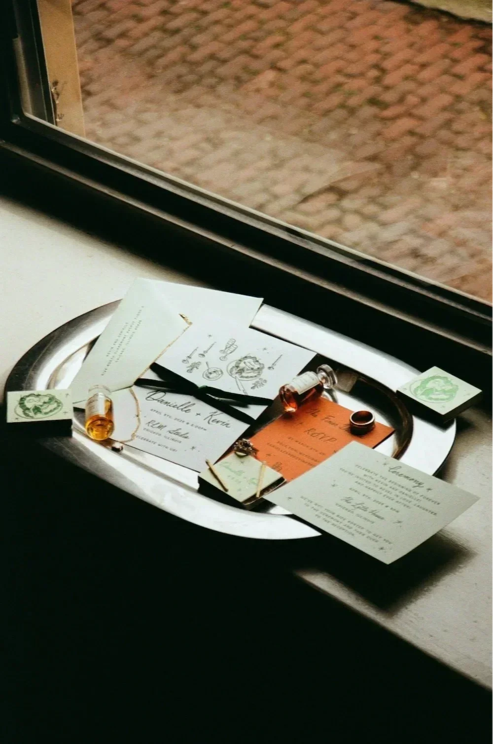 A silver tray holds wedding invitations and small bottles of essential oils or perfumes, placed on a windowsill with a view of a brick pathway outside.