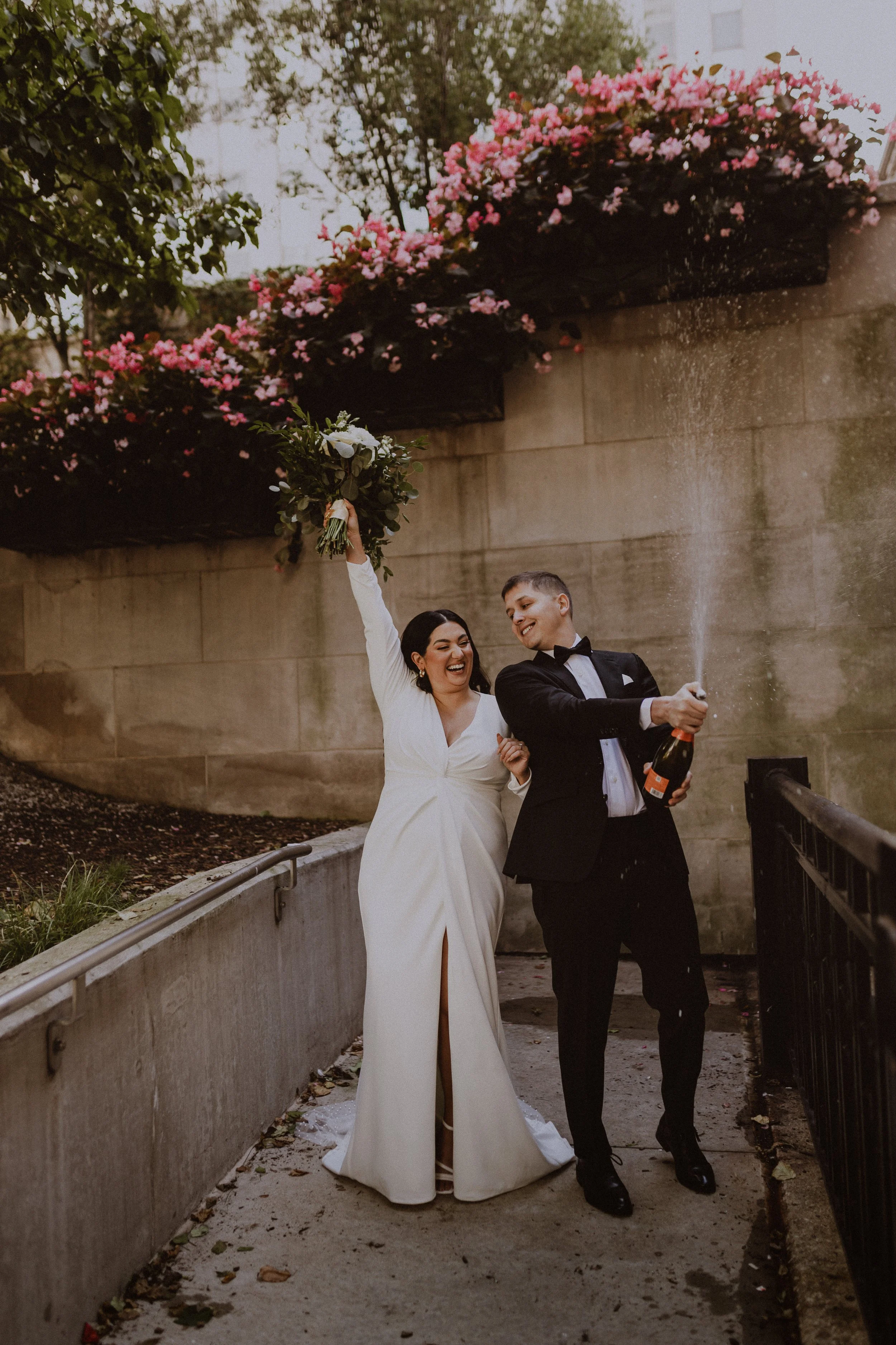 Happy newlywed couple celebrating with champagne outdoors, woman in white dress holding bouquet, man in tuxedo, pink flowers above them.