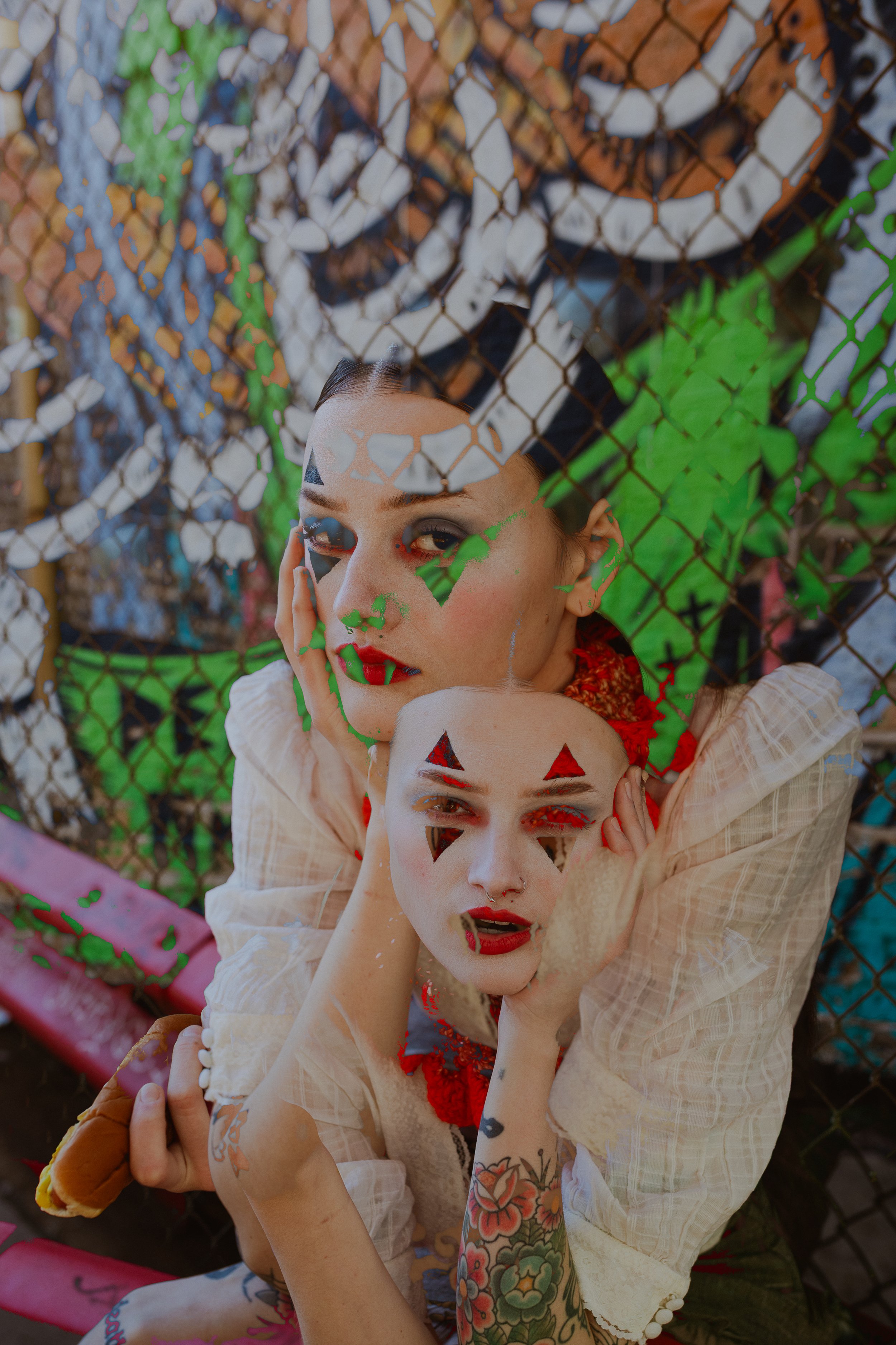 A non binary model, face paint poses in front of a graffiti wall. A double exposure creative portrait session with TTOLBPHOTOG- They have black triangles around the eyes, red lipstick, and facial piercings. They have tattoos and colorful clothing.
