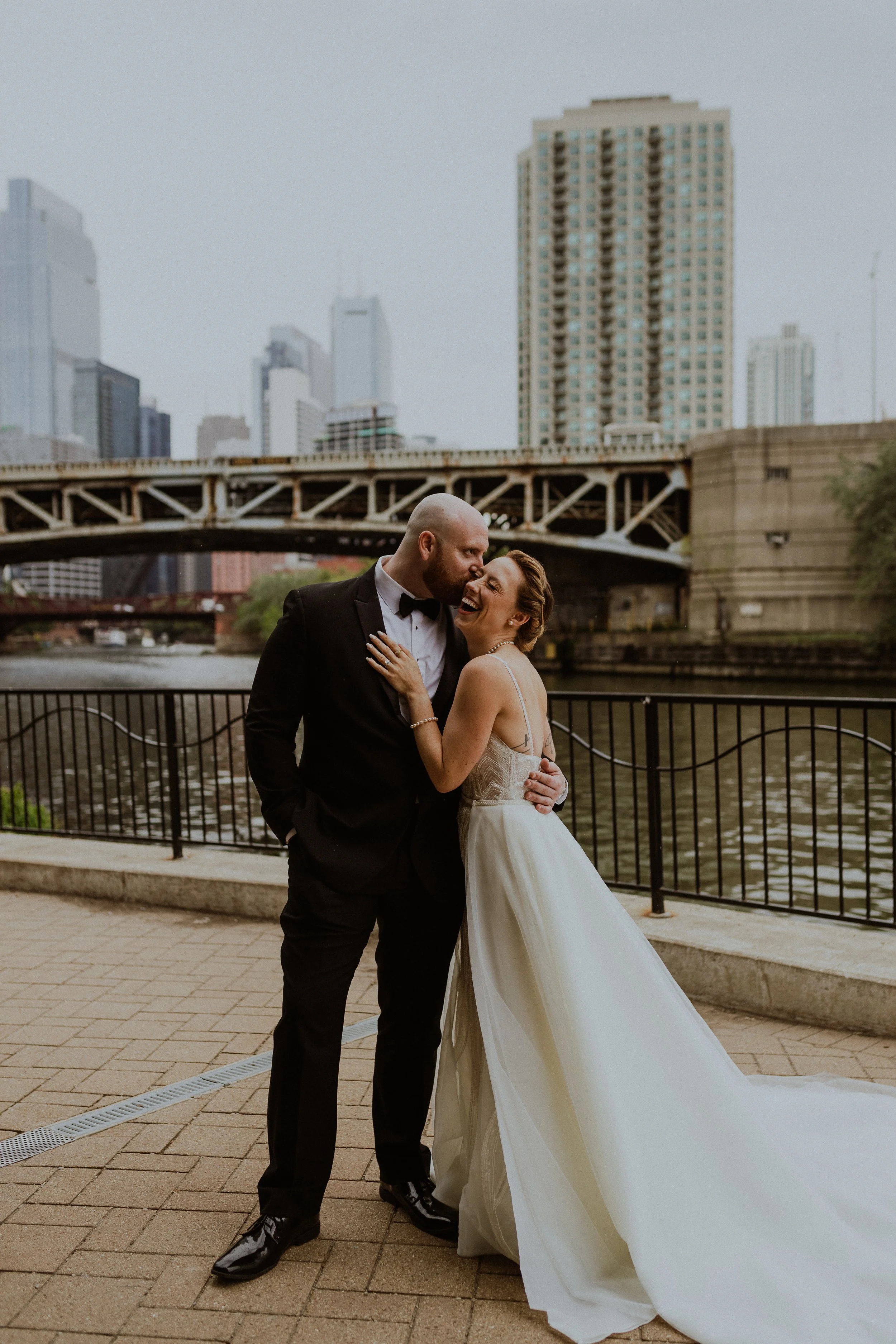 A newlywed couple dressed in formal wedding attire, embracing and smiling by a riverwalk with city buildings and a bridge in the background.