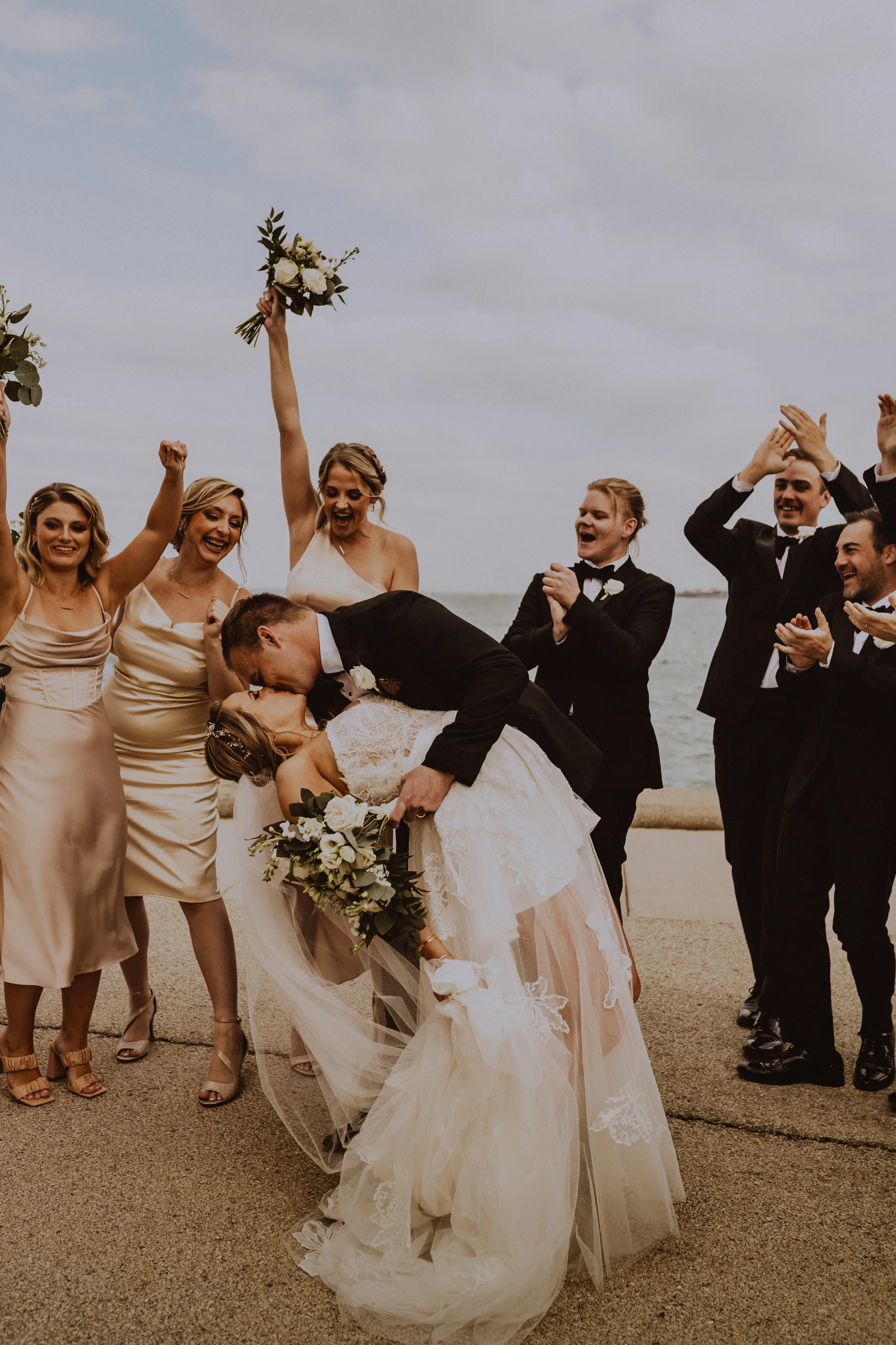 A bride and groom kiss as the bride leans backward during their wedding celebration by the water, surrounded by joyful friends and family clapping and cheering.