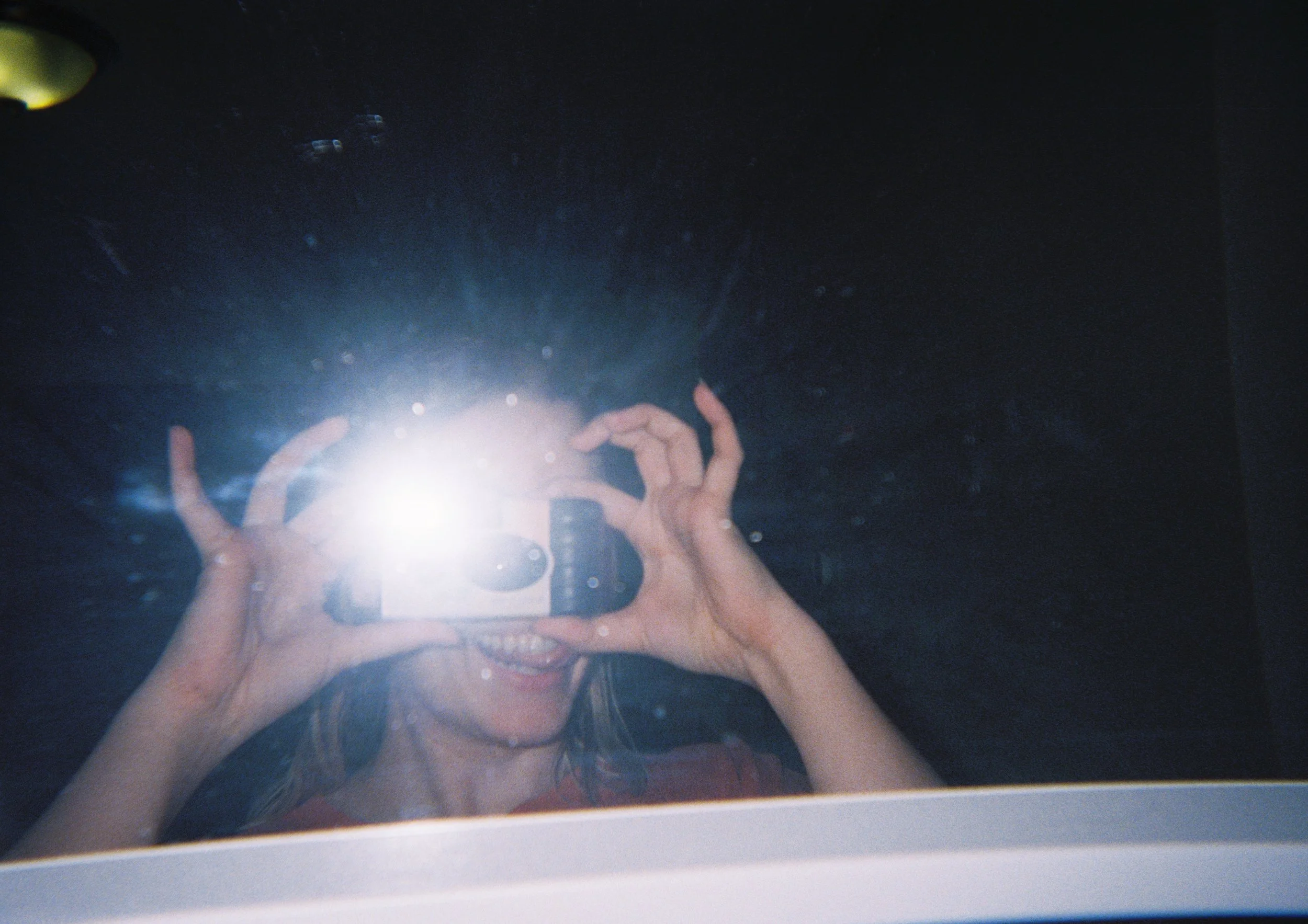 A person taking a selfie in a mirror, with the camera flash obscuring their face, showing a smile and holding the camera with both hands. The reflection appears to be in a dark room.