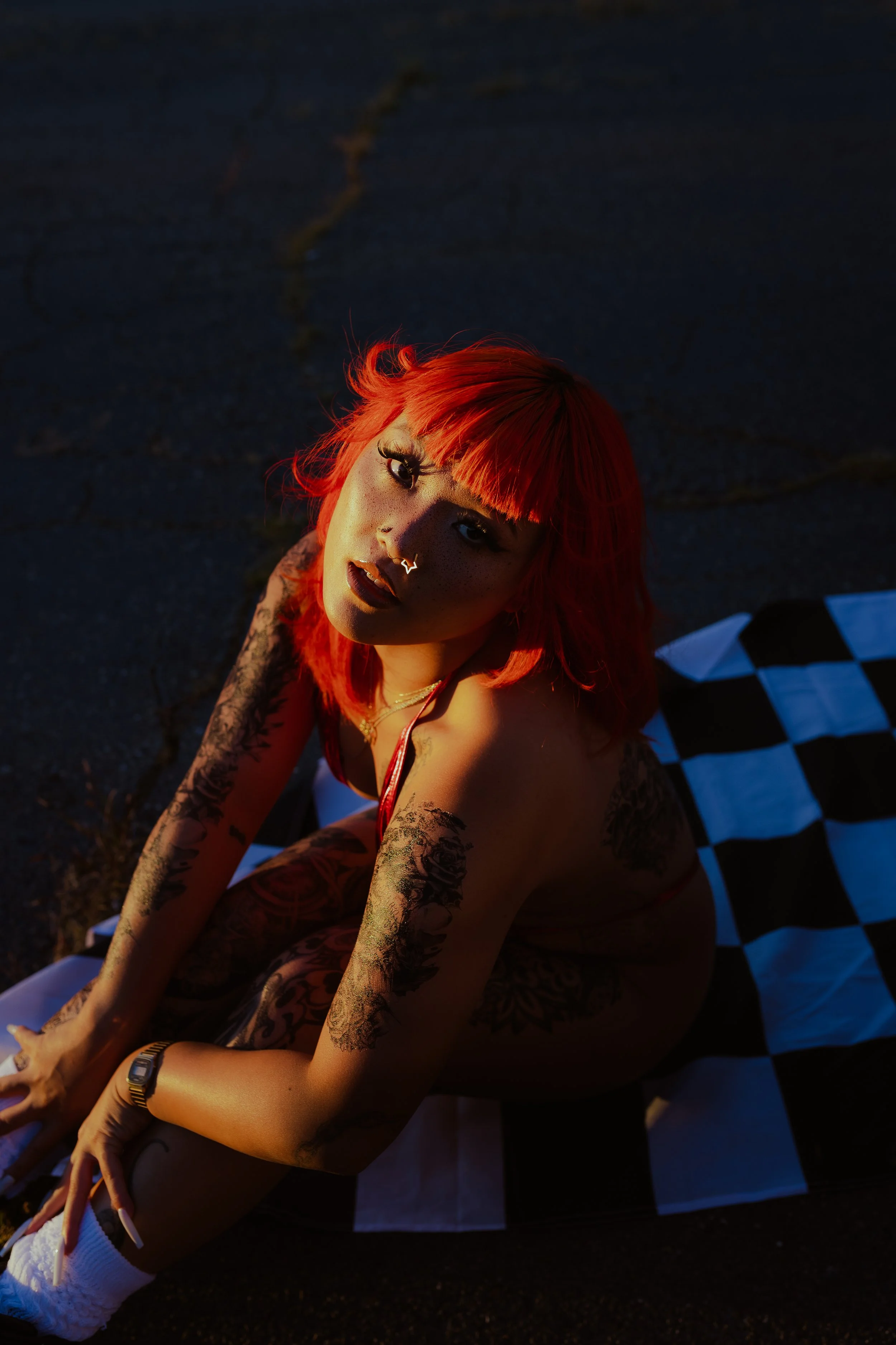 Ameya Okamoto pictured  in red bikini sitting on a checkered nascar flag with reddish orange hair flipped at ends with real tattoos and temporary tattoos showing at Sunset.