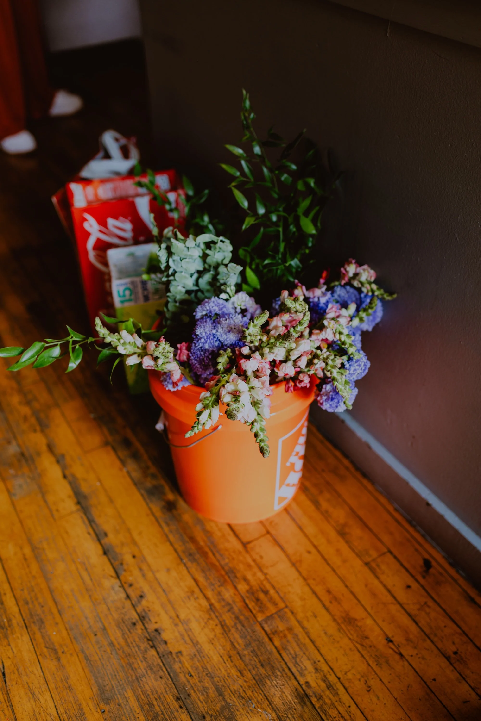 A flower arrangement in an orange pot contains purple, pink, and white flowers along with green leaves, placed on a wooden floor next to a gray wall.