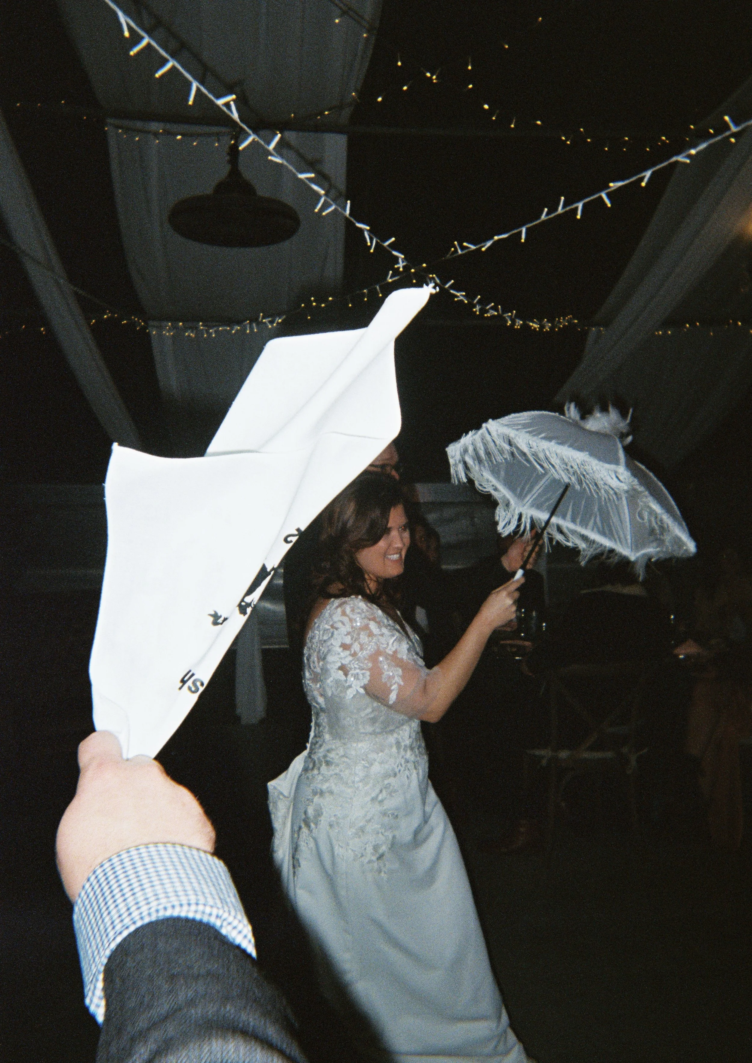 A woman in a wedding dress holding a white umbrella, smiling, at a celebration with string lights and décor overhead, and a hand in the foreground holding an open white umbrella.