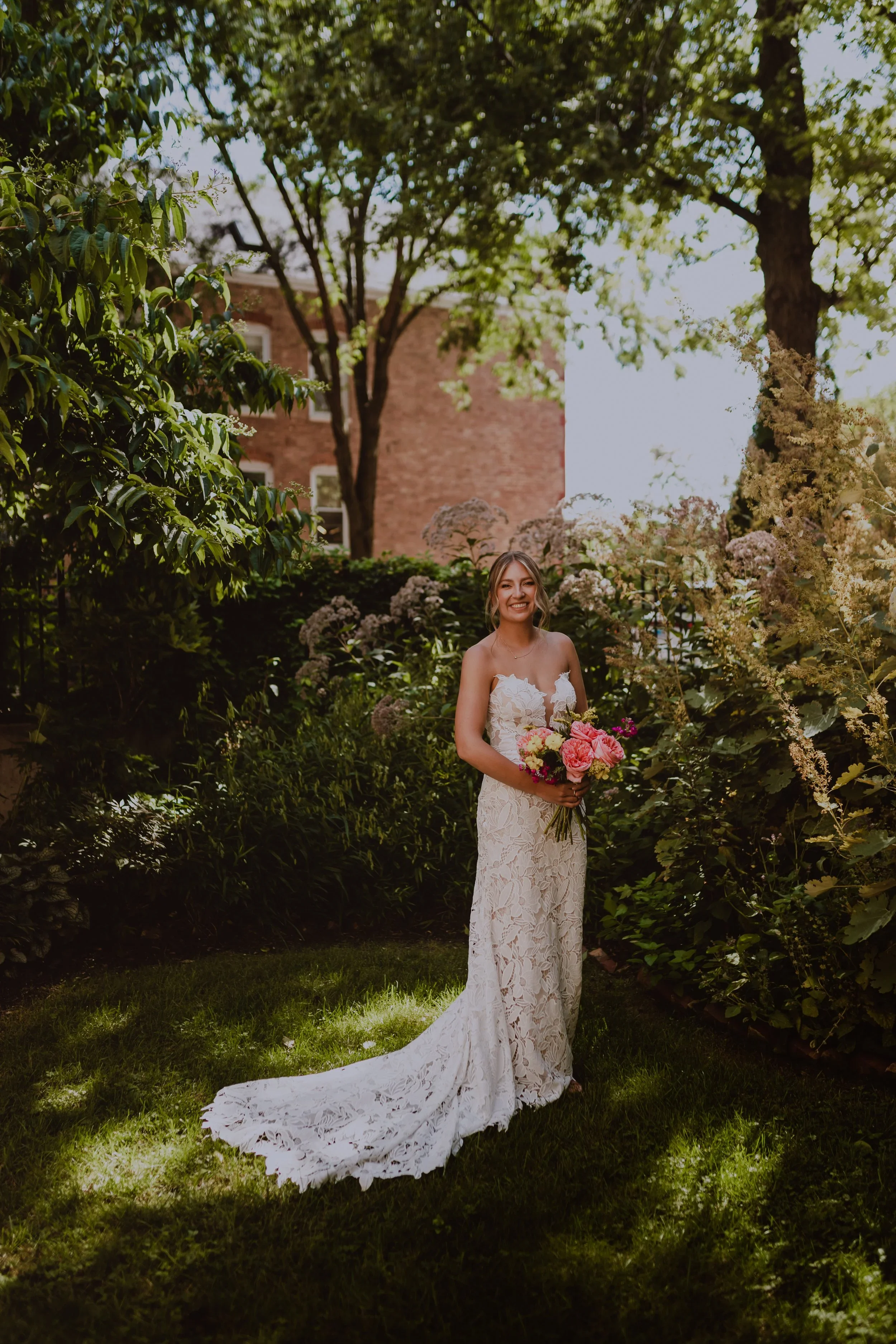 A Bride in a white lace wedding dress holding a bouquet of pink and white flowers, standing in a lush garden with trees and bushes, smiling. Wedding photography by ttolbphotog a Chicago based candid photographer 