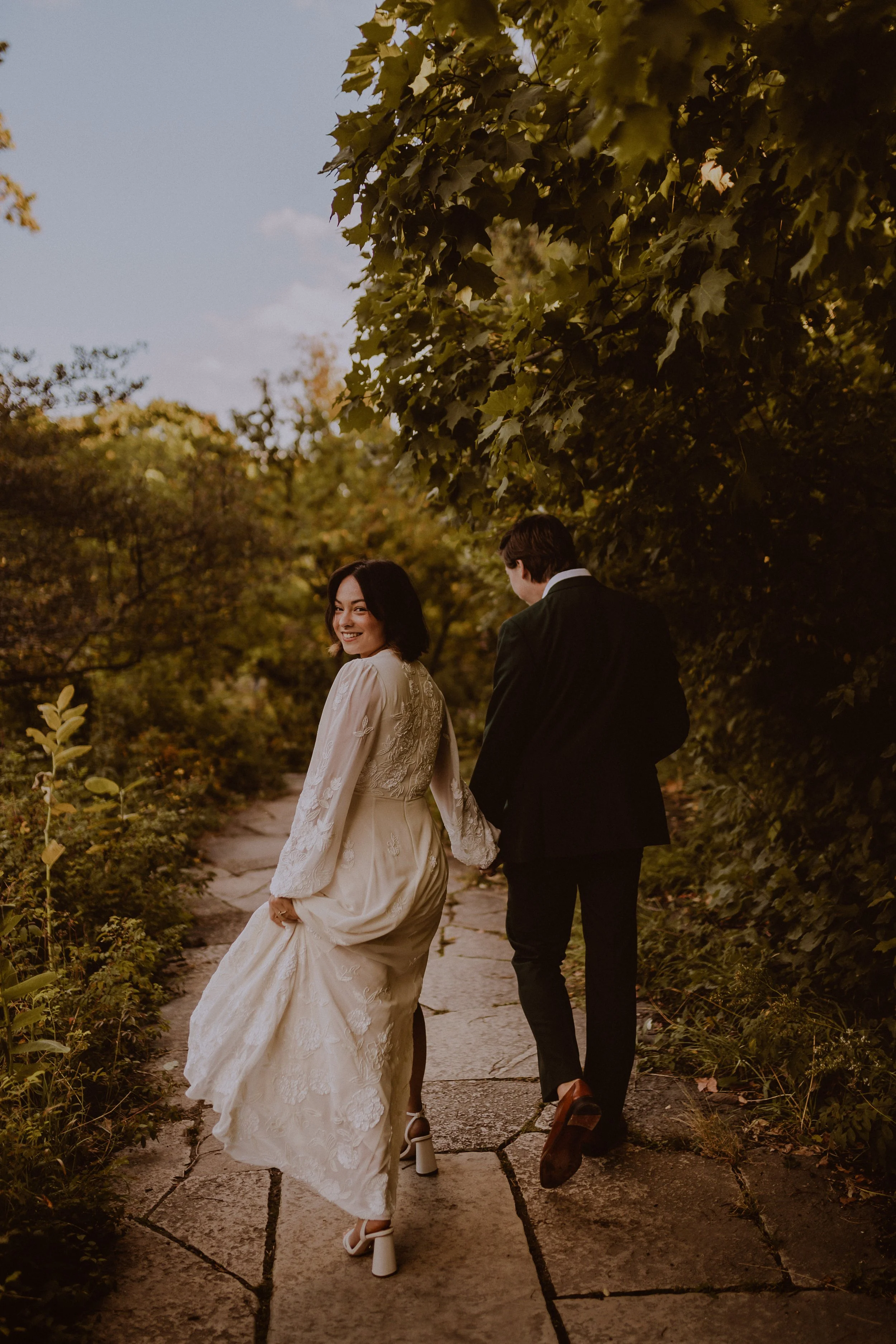 A couple walking on a stone pathway in a wooded area, holding hands. The woman is wearing a cream-colored dress and smiling while the man is dressed in a black suit, shown from behind. Alfred Caldwell Lilly Pool Lincoln Park Chicago