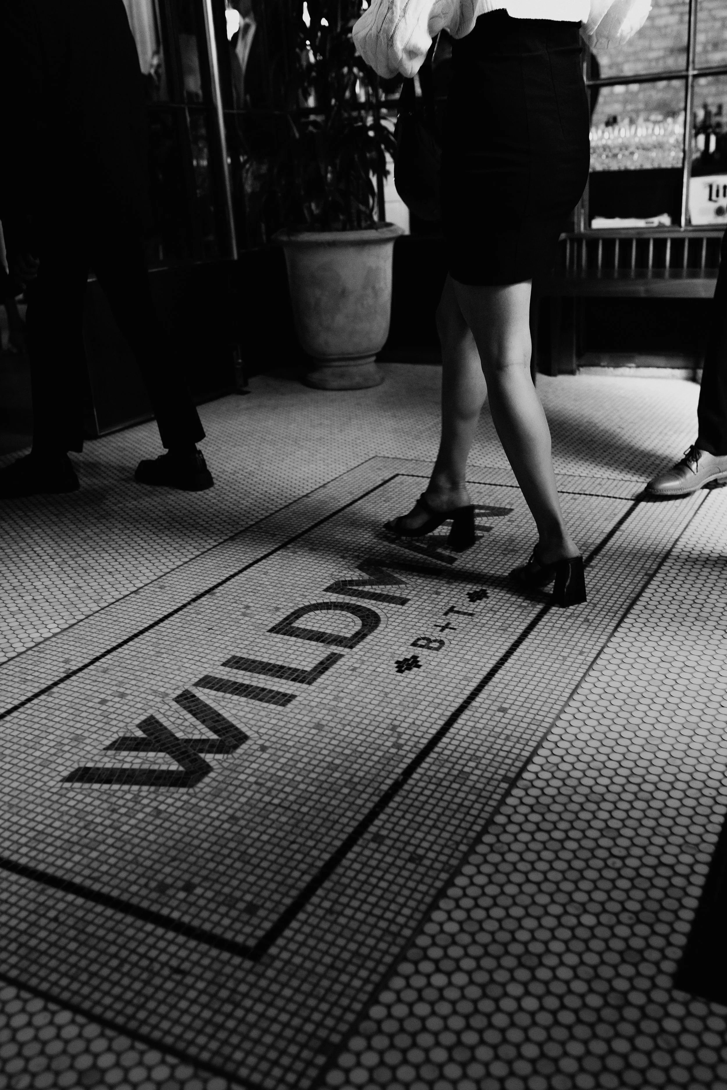 Black and white photo of a woman standing on a tiled floor with the word 'WILDE' and some numbers written on it. She is wearing high heels, a skirt, and a light-colored top, with her back turned to the camera. There are other people partially visible