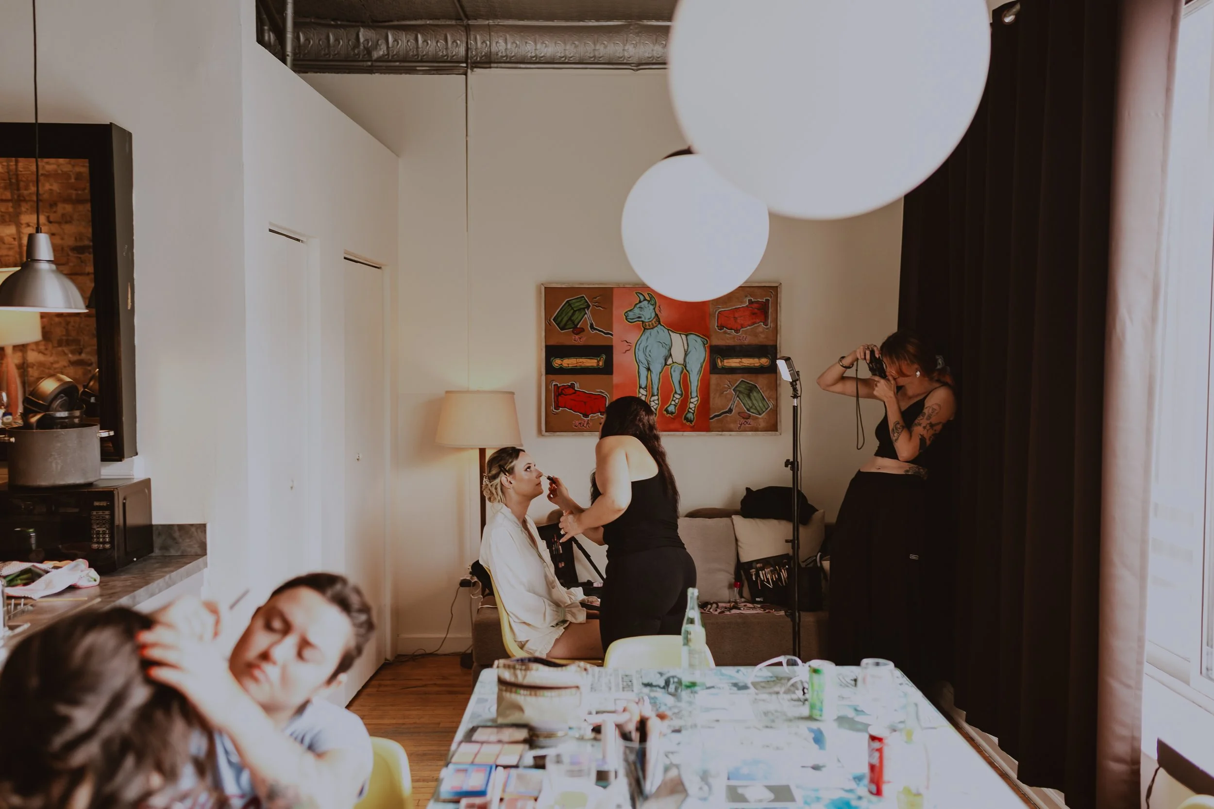 A woman sitting on a chair getting her makeup done by another woman in a living room. Another woman is taking a photo of the scene with a camera. There is a painting of a unicorn on the wall, and the table in the foreground has makeup and beauty prod
