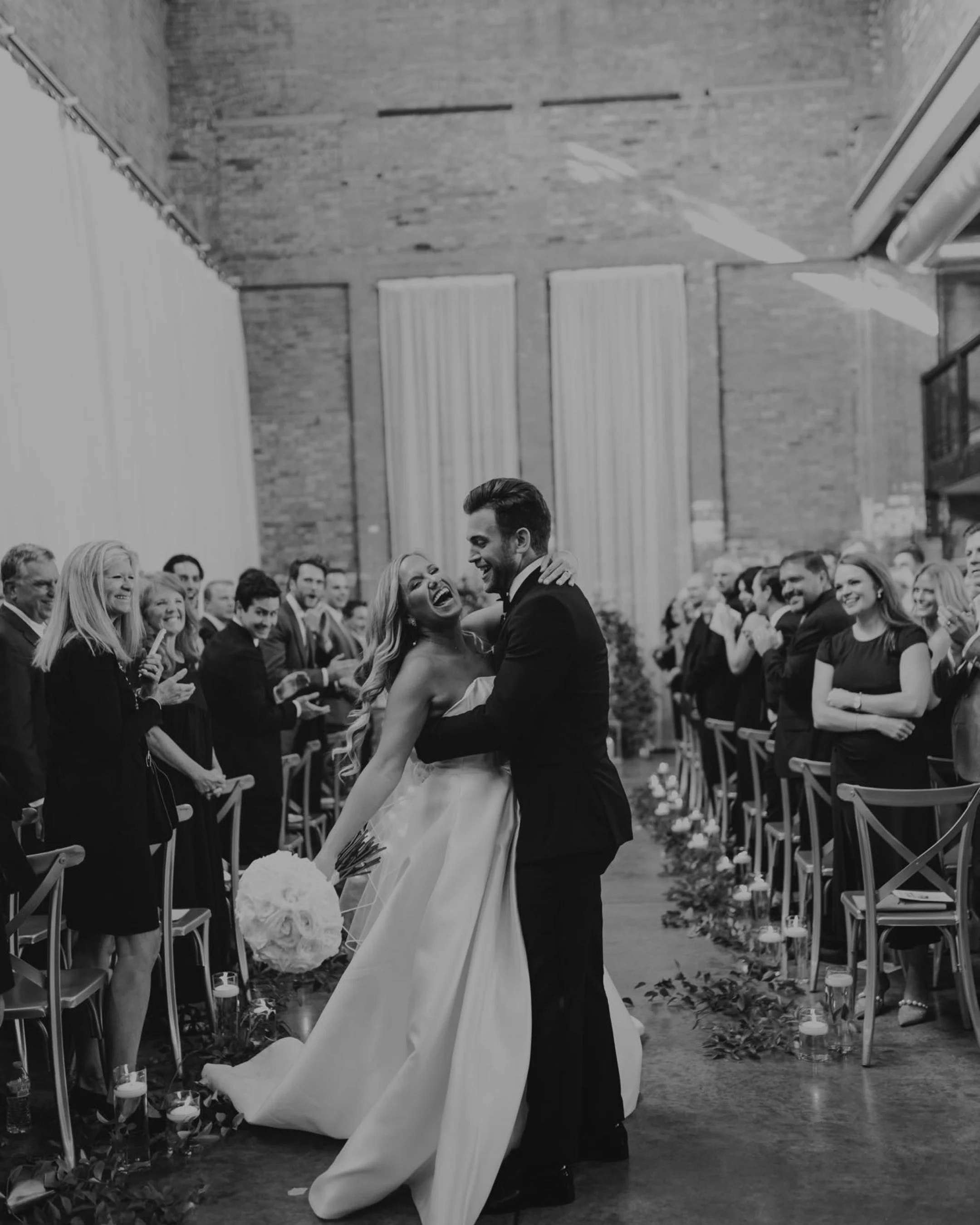 A bride and groom dancing and laughing at their wedding ceremony, surrounded by smiling guests in an indoor venue.