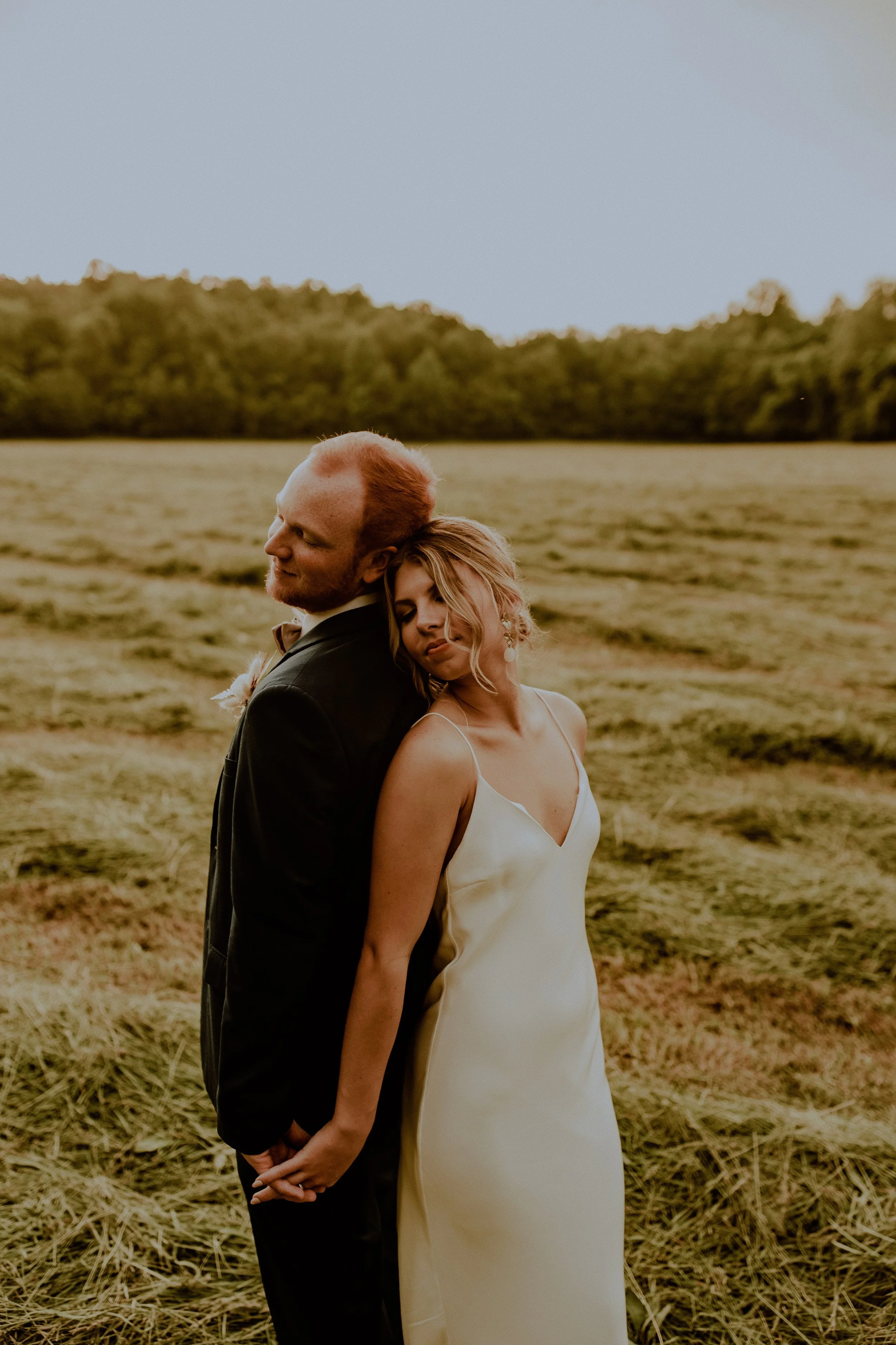 A bride and groom holding hands and standing back to back in a field at sunset with trees in the background.