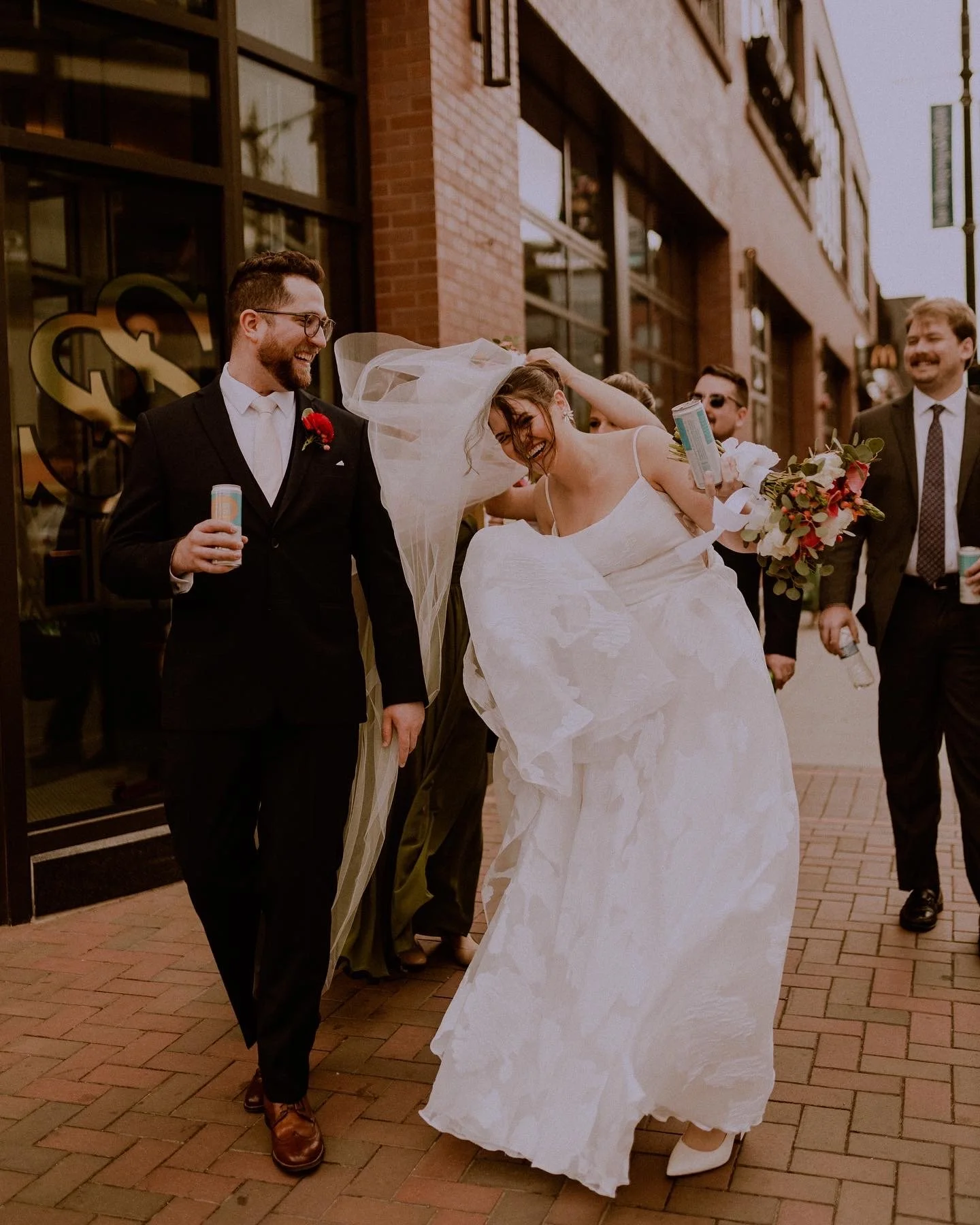 A bride and groom are surrounded by friends on a city sidewalk, celebrating their wedding as they smile and laugh. The bride is in a white wedding dress and veil, while the groom is in a black suit with a red boutonniere. Friends are holding drinks a