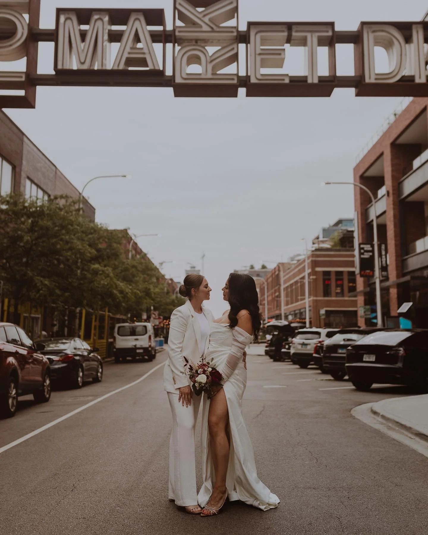 Two women in wedding attire standing close together on a city street, gazing at each other, with one holding a bouquet of flowers. Buildings and cars are visible in the background, under an overcast sky.