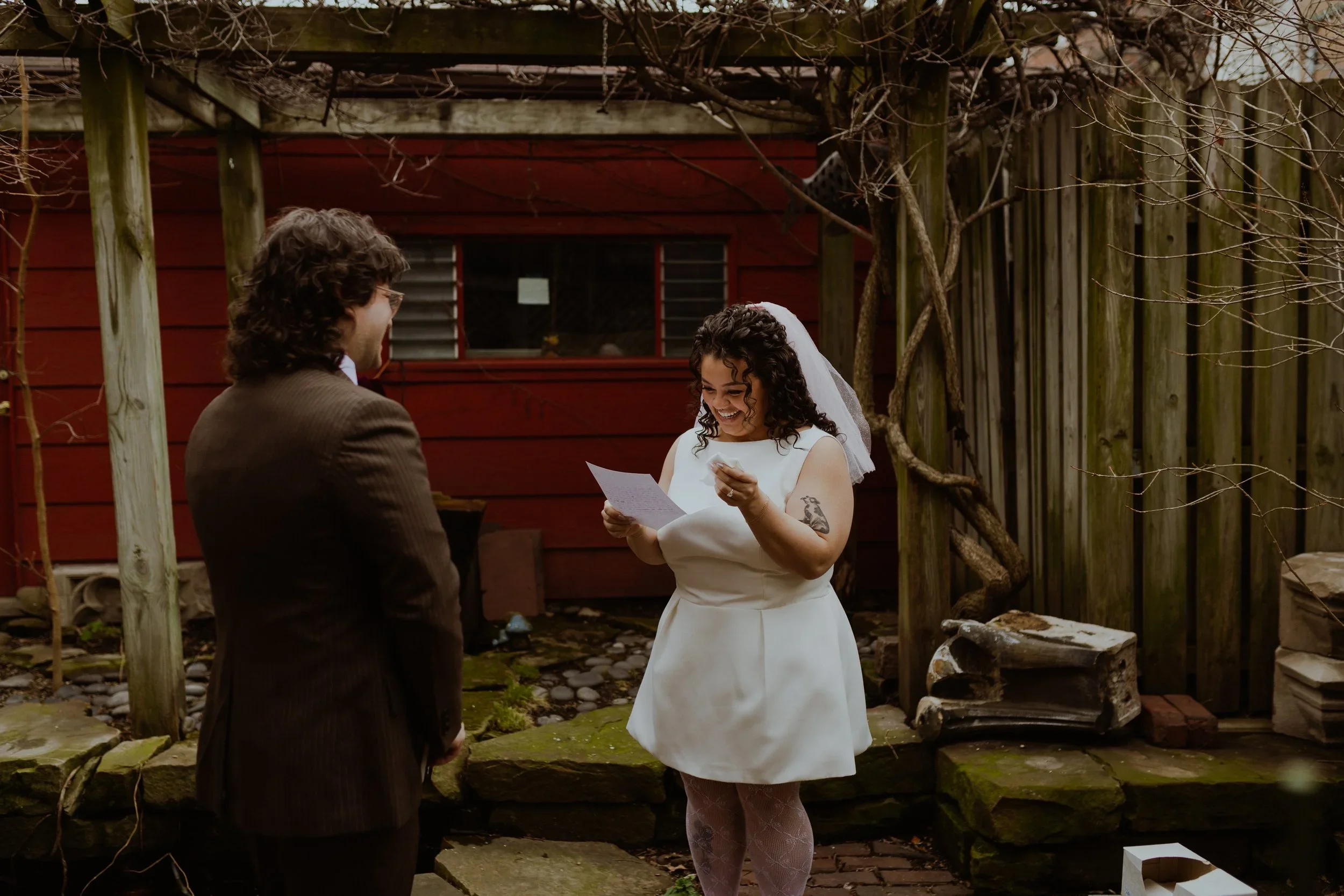 A woman in a white dress and veil, holding a paper and reading it, is smiling and looking at a man in a brown suit, who is facing her and listening. They are standing outdoors in a backyard with a red wooden shed and a wooden fence.