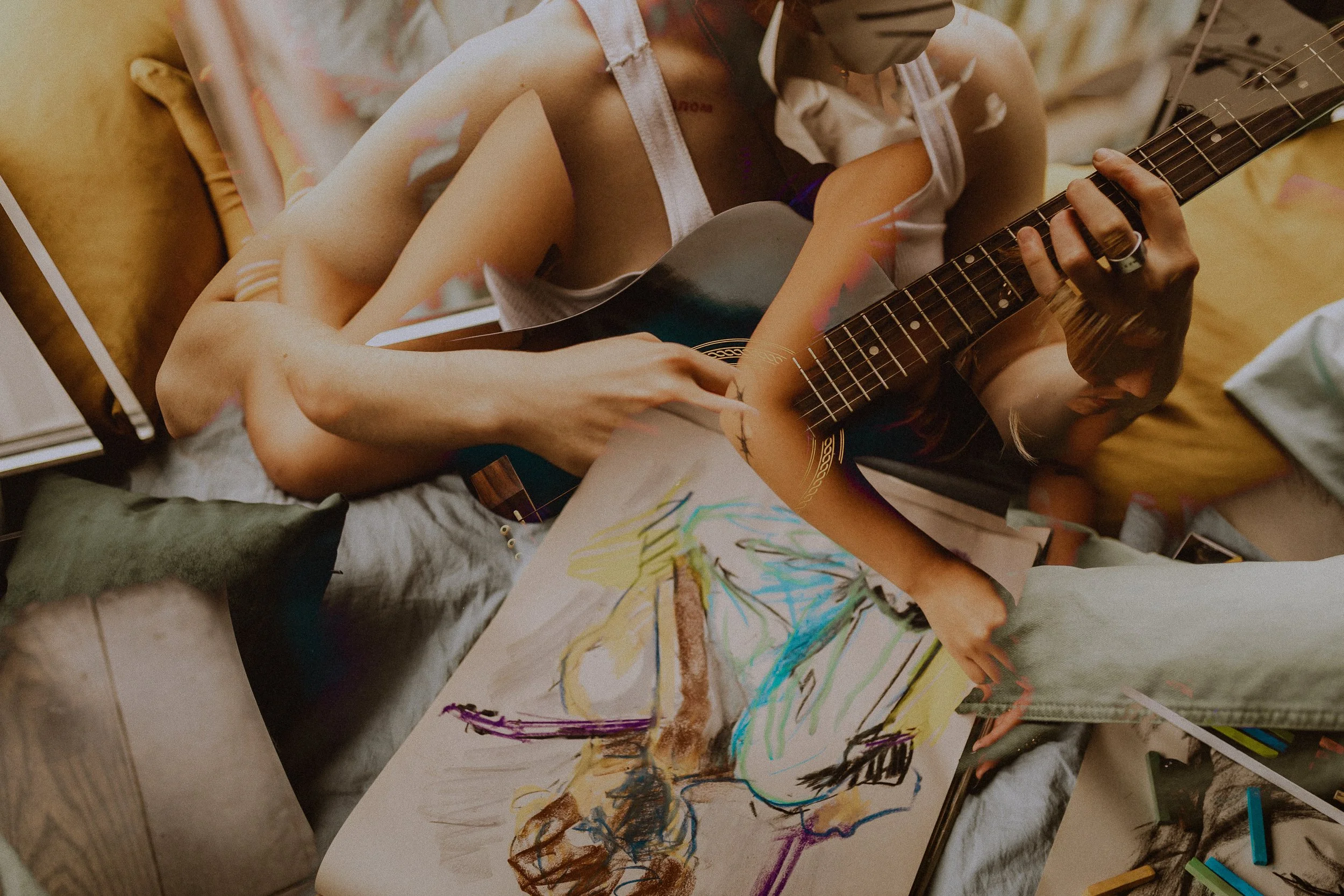 A man playing an acoustic guitar while a woman lying on a bed with colorful artwork and art supplies around her. Double exposure by ttolbphotog