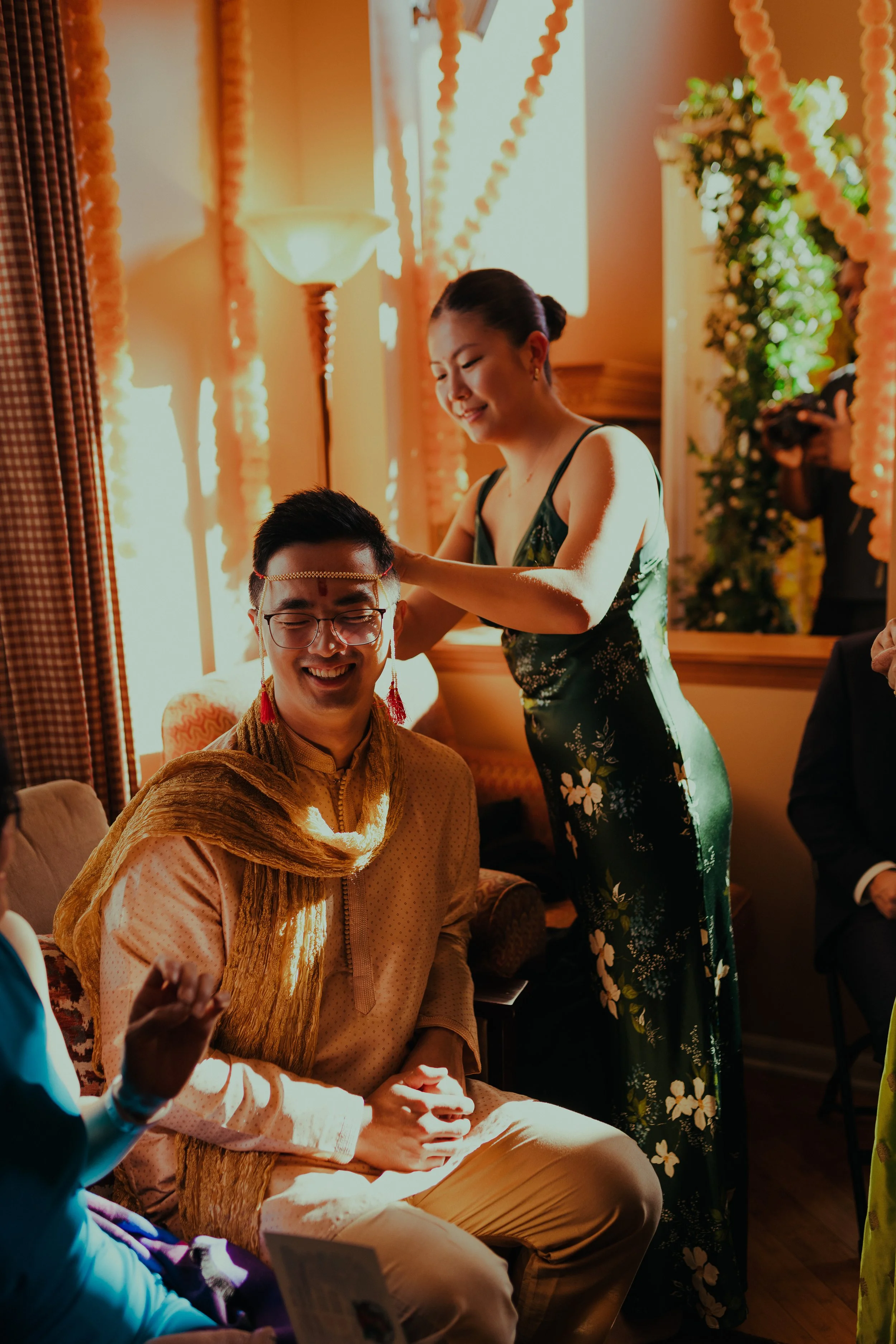 A woman in a black floral dress places a necklace on a smiling man wearing glasses and traditional Indian attire during a celebration.