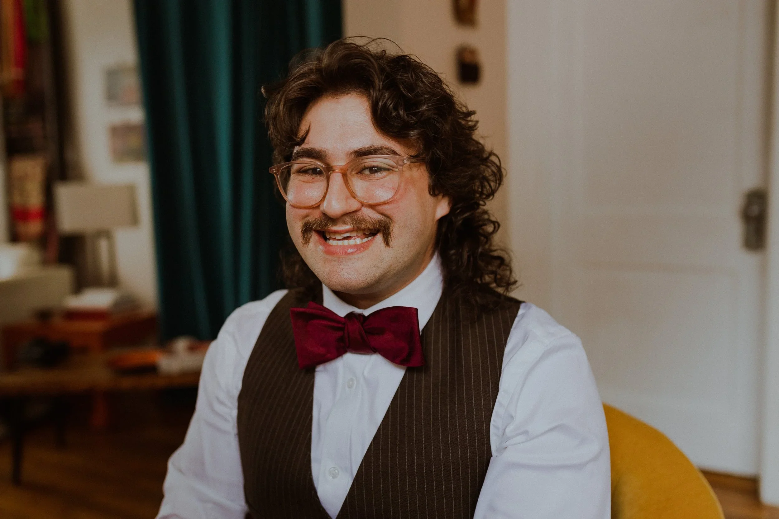 A man with curly brown hair, glasses, and a mustache, wearing a white dress shirt, dark striped vest, and red bow tie, smiling in a room with curtains and books in the background.