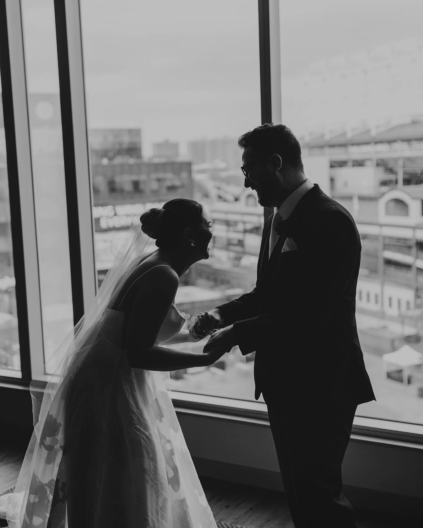 Black and white photo of a bride and groom holding hands, laughing and facing each other in front of large windows with an urban cityscape in the background.