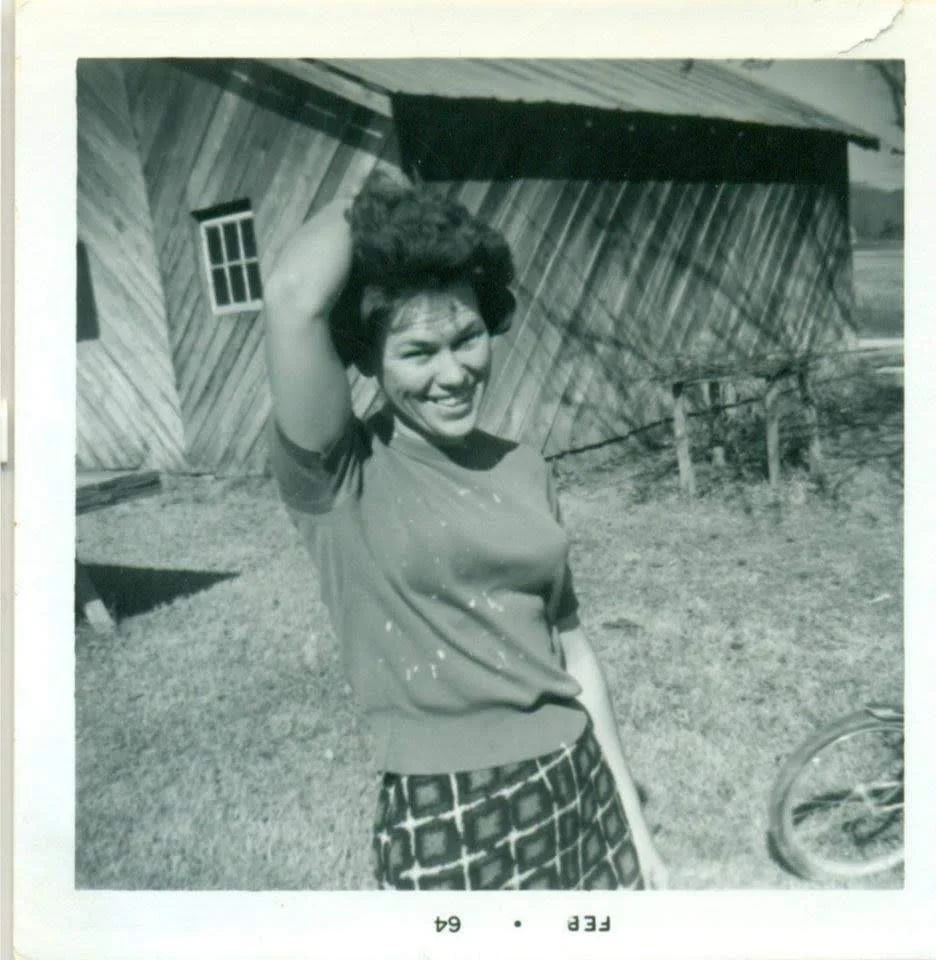 A woman smiling and posing outdoors in front of a wooden building, with one hand raised behind her head and the other by her side, wearing a short-sleeved shirt and patterned pants.
