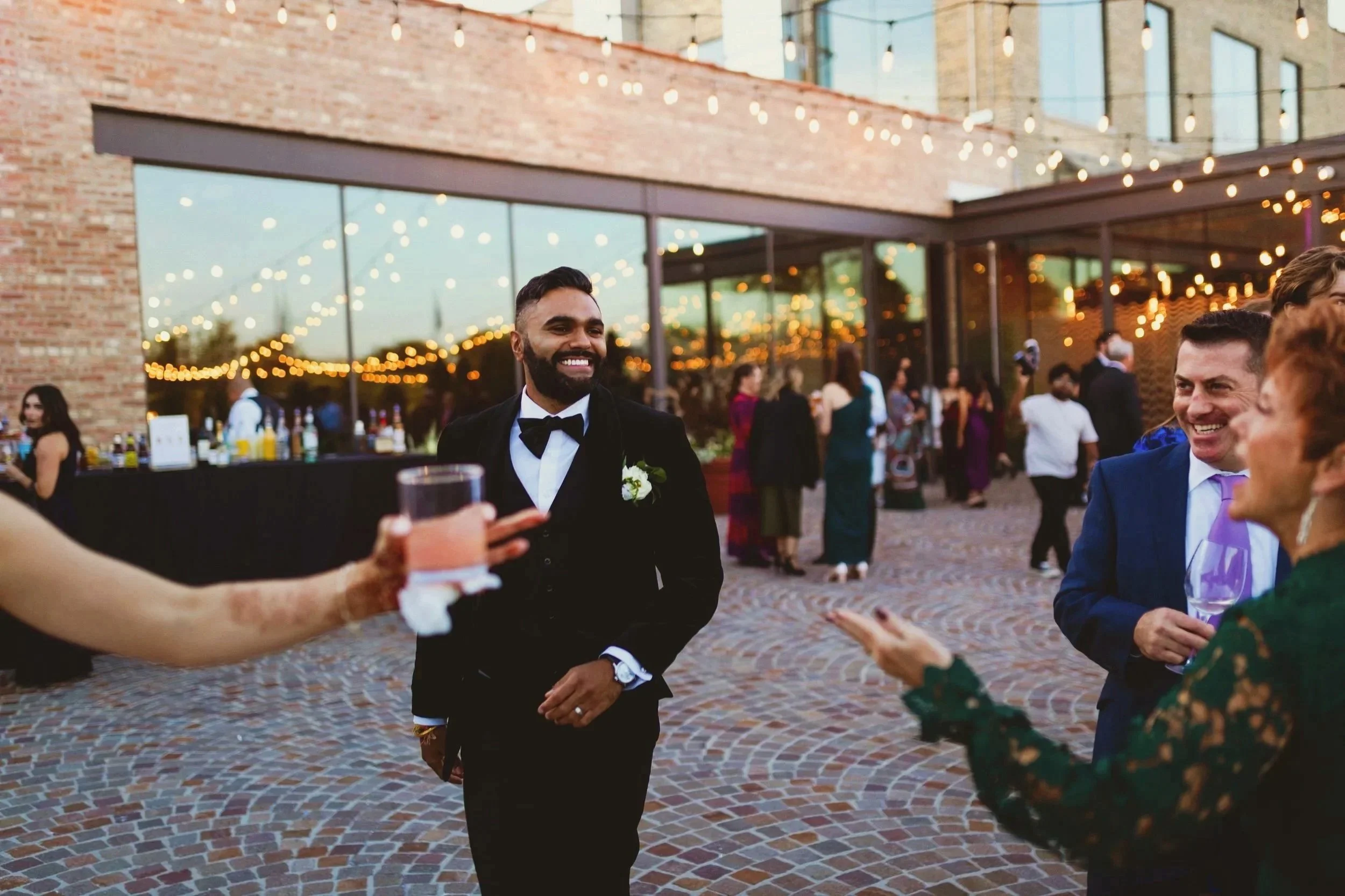 People at a wedding reception socializing outdoors at Rockwell On The River, Groom in a black tuxedo smiling in the foreground, and others holding drinks in a festive atmosphere with string lights and a modern brick building in the background.