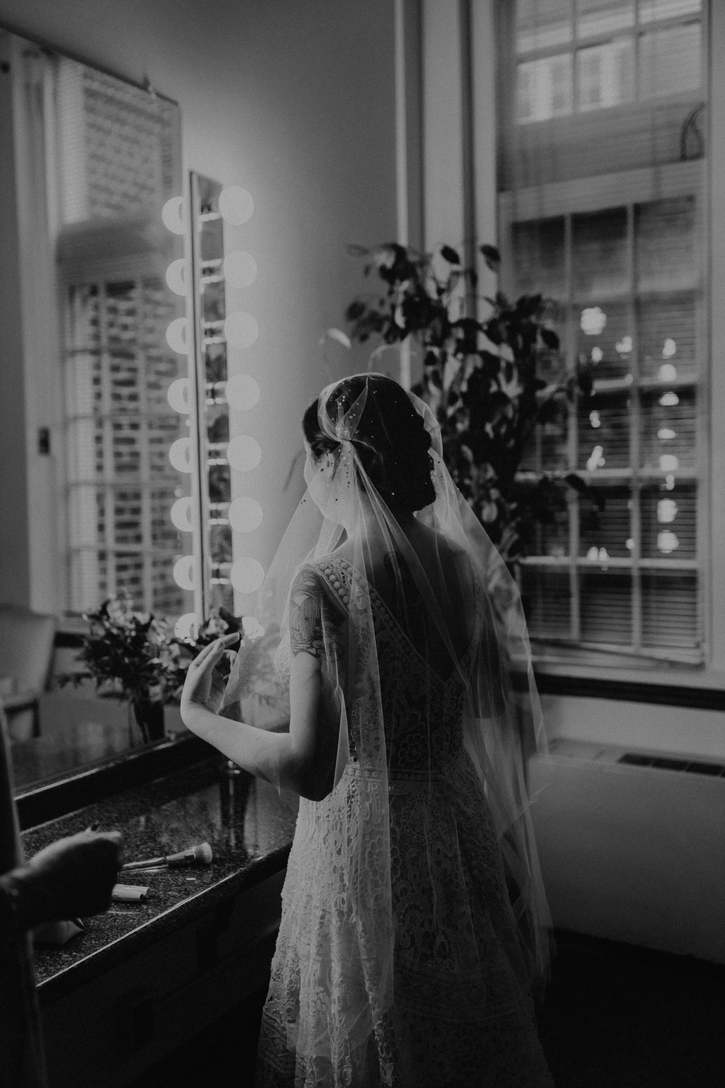 A bride standing in front of a mirror with lights, wearing a veil and lace dress, with tattoos on her arm, in a room with a window and a plant.