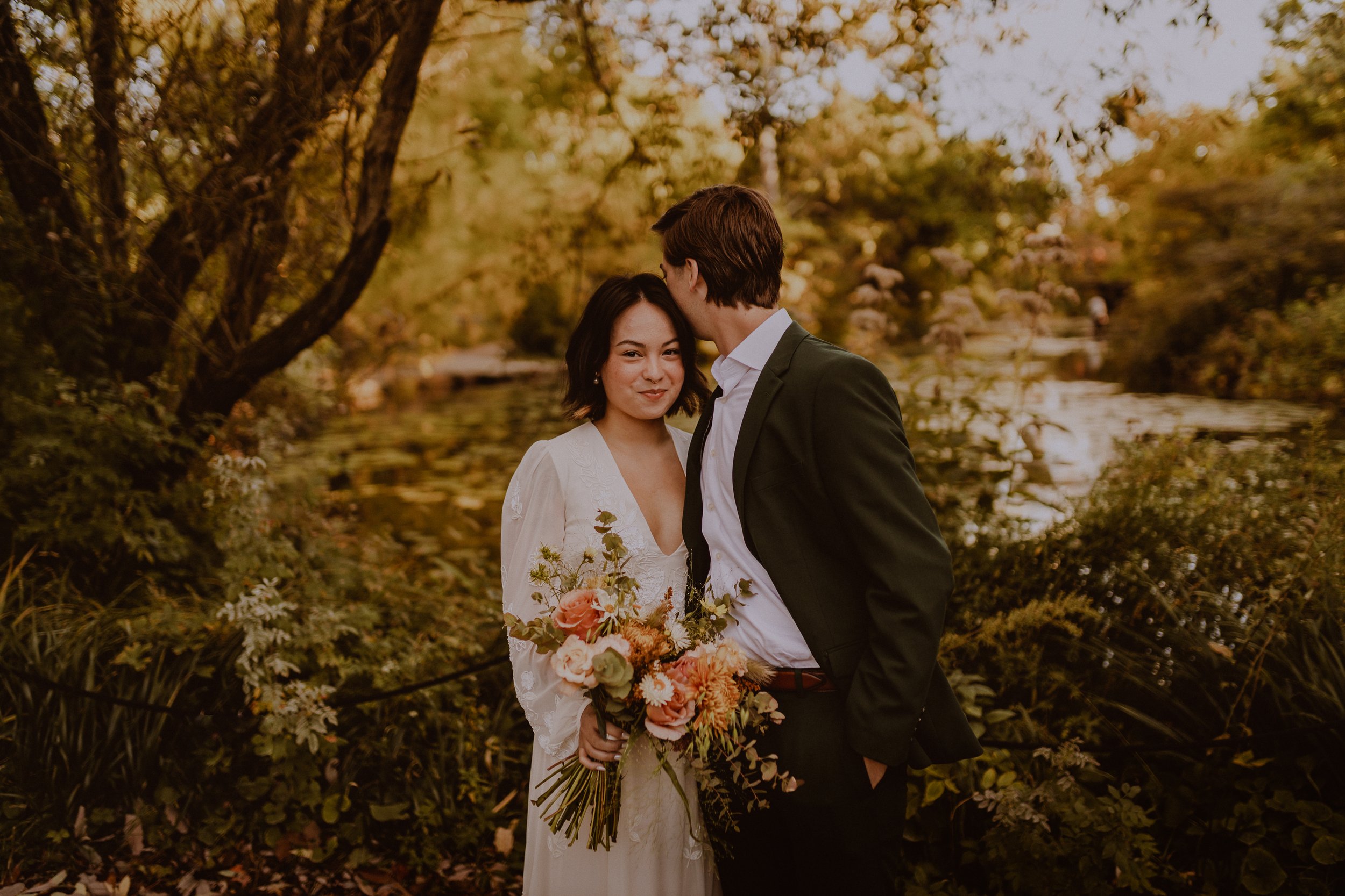A couple dressed in wedding attire standing outdoors near a pond, with the woman holding a bouquet of flowers and the man leaning in to kiss her on the forehead, surrounded by trees with autumn-colored leaves.