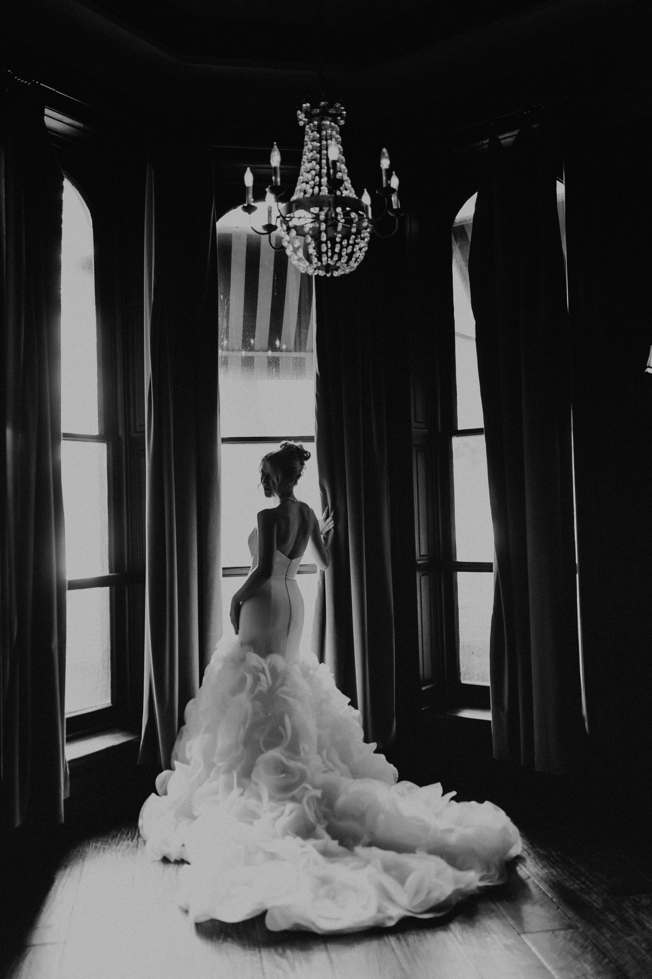 A bride in a strapless wedding gown with a voluminous, ruffled train standing by large windows with curtains in a dark room, illuminated by natural light and a chandelier overhead.