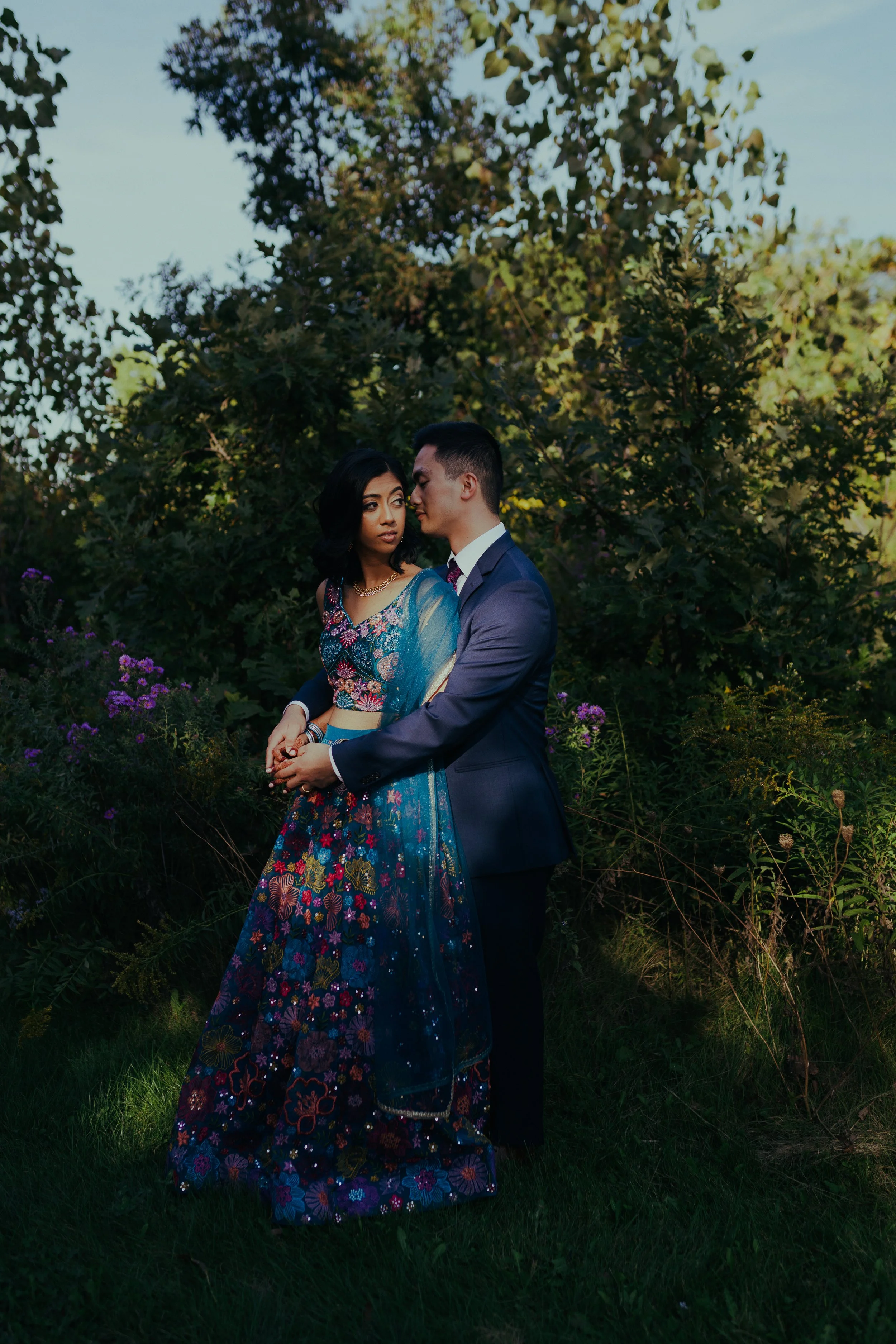 A couple standing outdoors among trees and purple flowers, dressed in colorful, formal attire, gazing at each other affectionately.