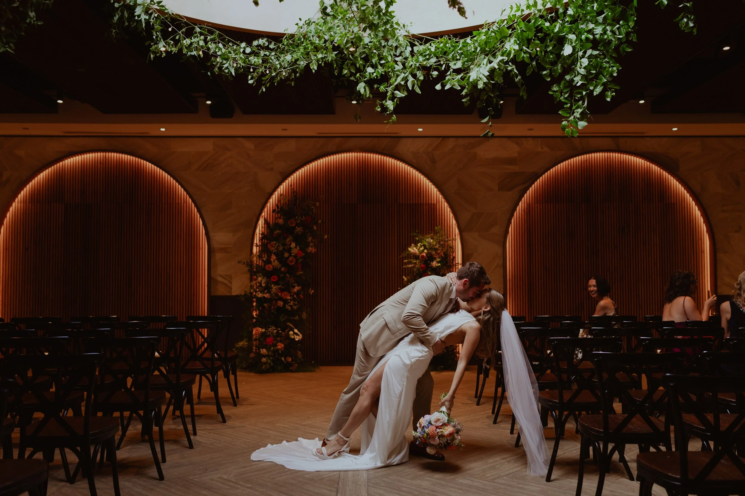 A newlywed couple sharing a romantic kiss during their wedding reception in a decorated indoor venue, with friends seated at tables in the background.