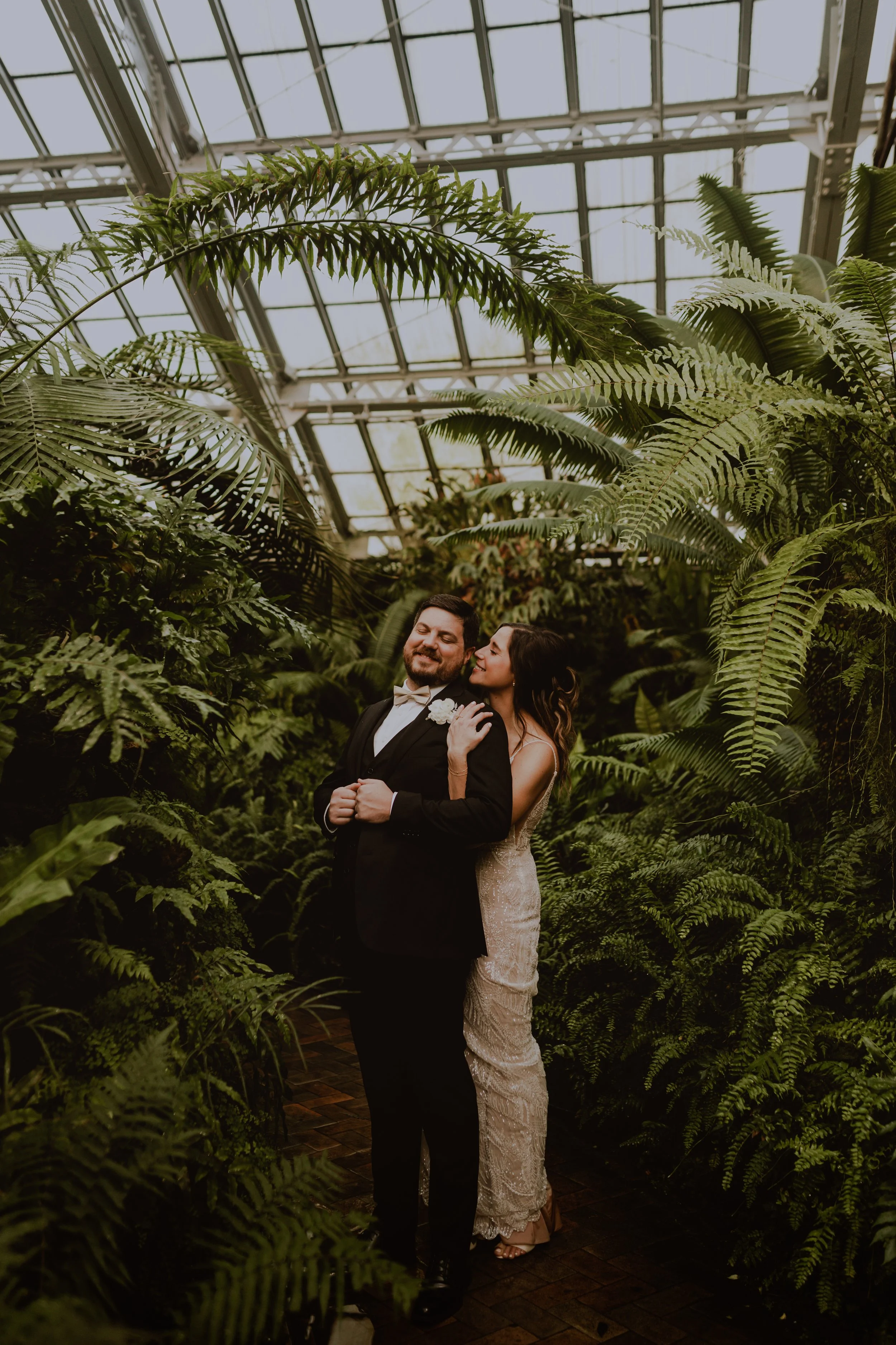 A bride and groom standing in front of lush green plants and ferns inside a greenhouse, smiling and embracing each other.