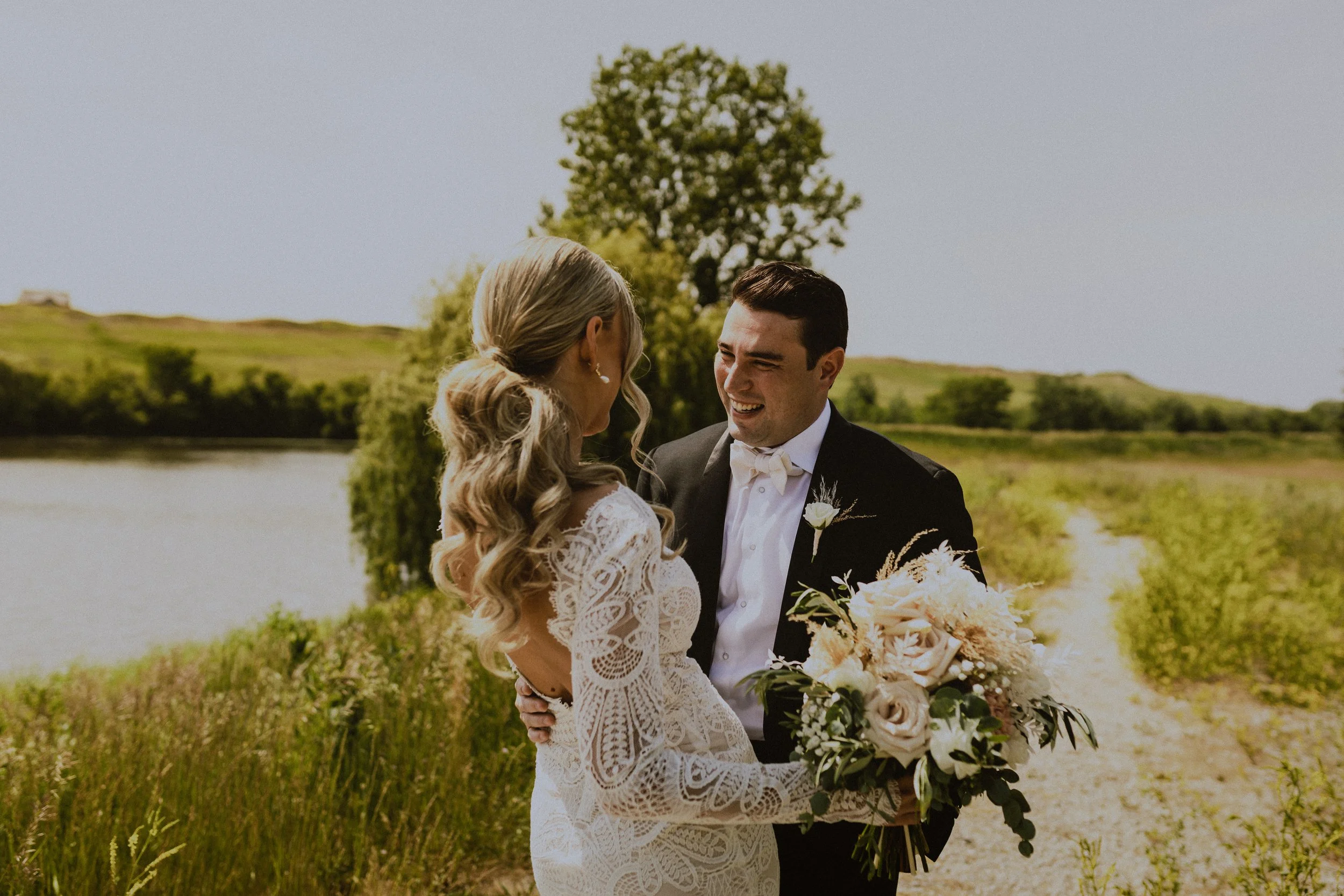 A bride and groom smiling at each other outdoors near a body of water, with greenery and a tree in the background during a wedding.