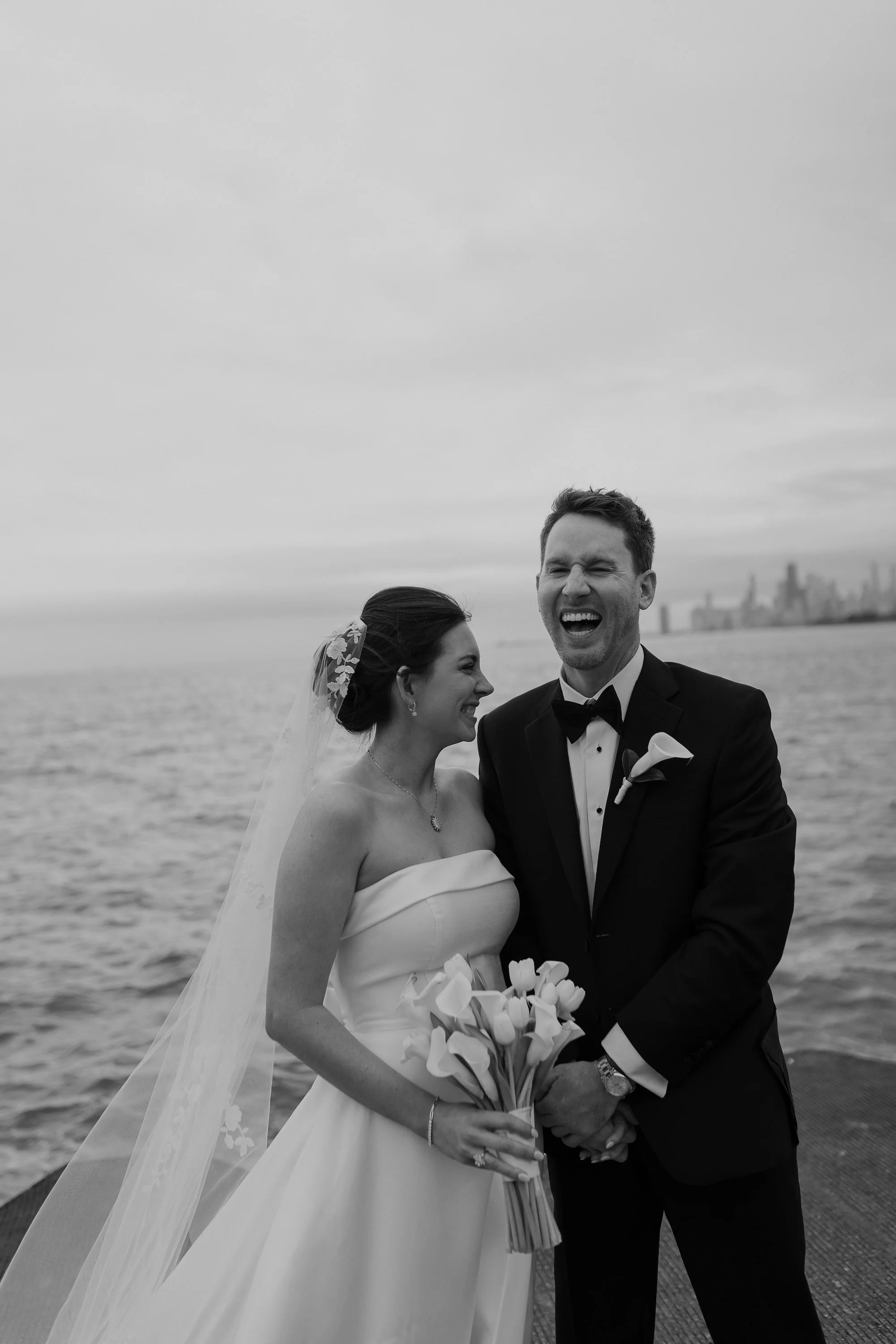 Black and white photo of a newlywed couple laughing and holding a bouquet of calla lilies near the water with a Chicago city skyline in the background.