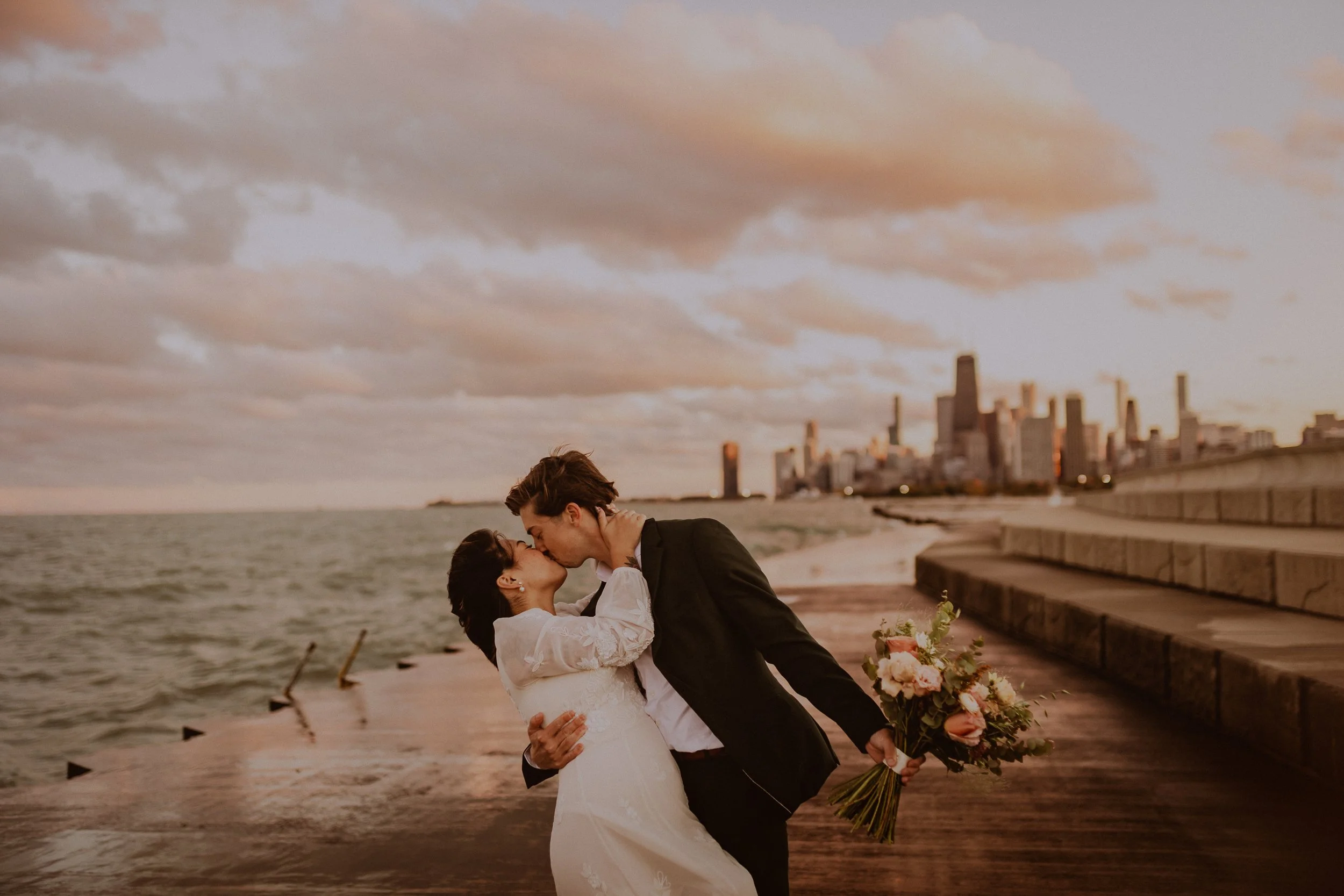 A bride and groom sharing a kiss on the Chicago lakefront with a city skyline in the background, the bride holding a bouquet of flowers. Elopement Photos by Ttolbphotog a Chicago Based Photographer