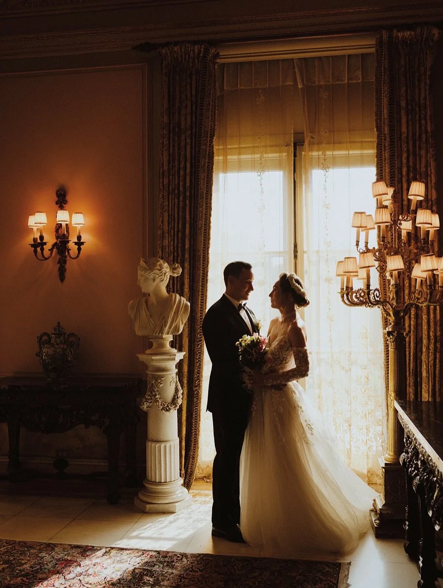 Silhouette of a bride in a white wedding dress holding a bouquet and groom in a tuxedo standing close together in a bright, elegant room with classical decor, lace curtains, statues, and ornate wall sconces.