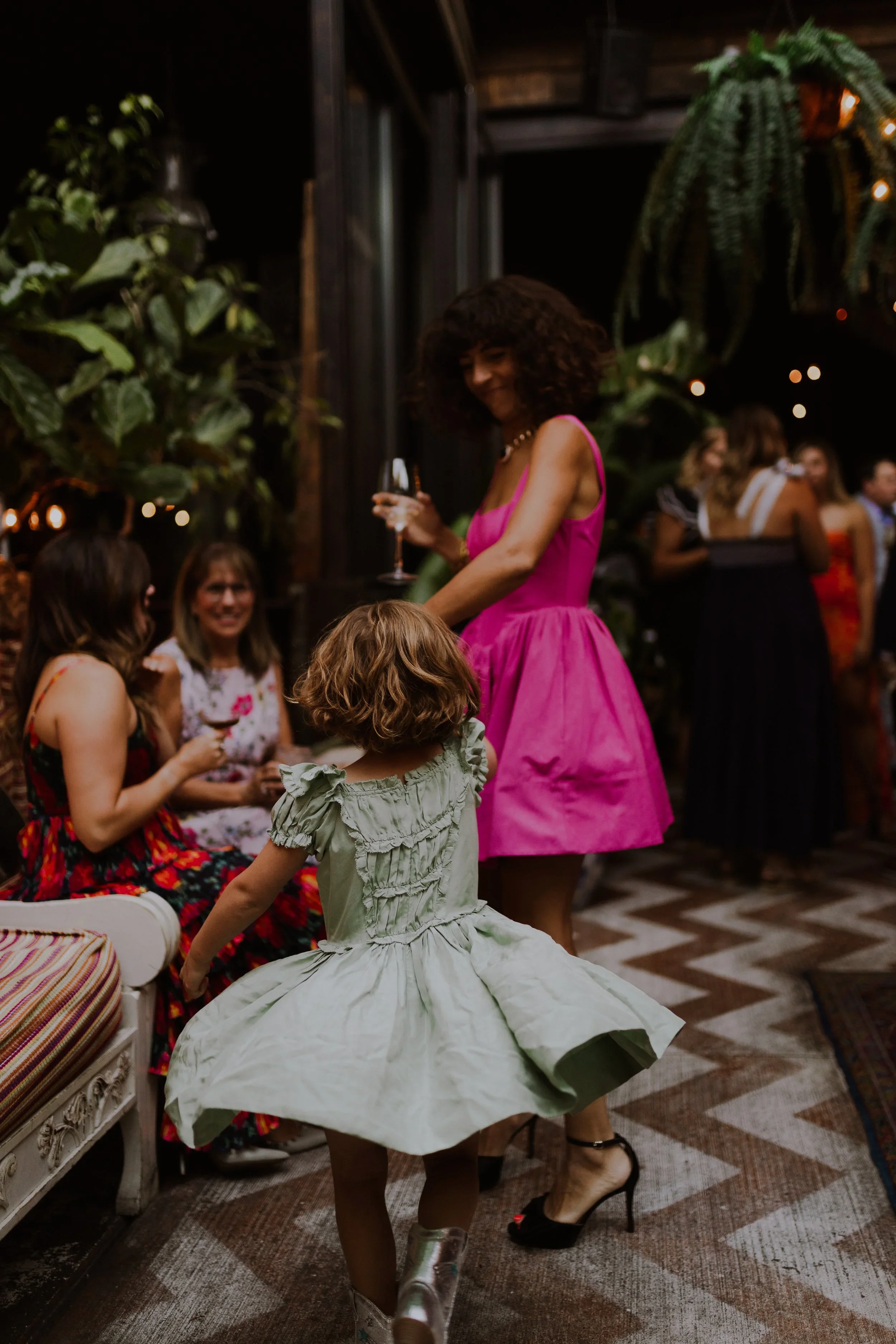 A woman in a pink dress holding a glass and smiling at a young girl with curly hair wearing a light green dress at a Wedding Reception gathering indoors at Beatnik West Town with multiple people in the background. Intimate Wedding Images 