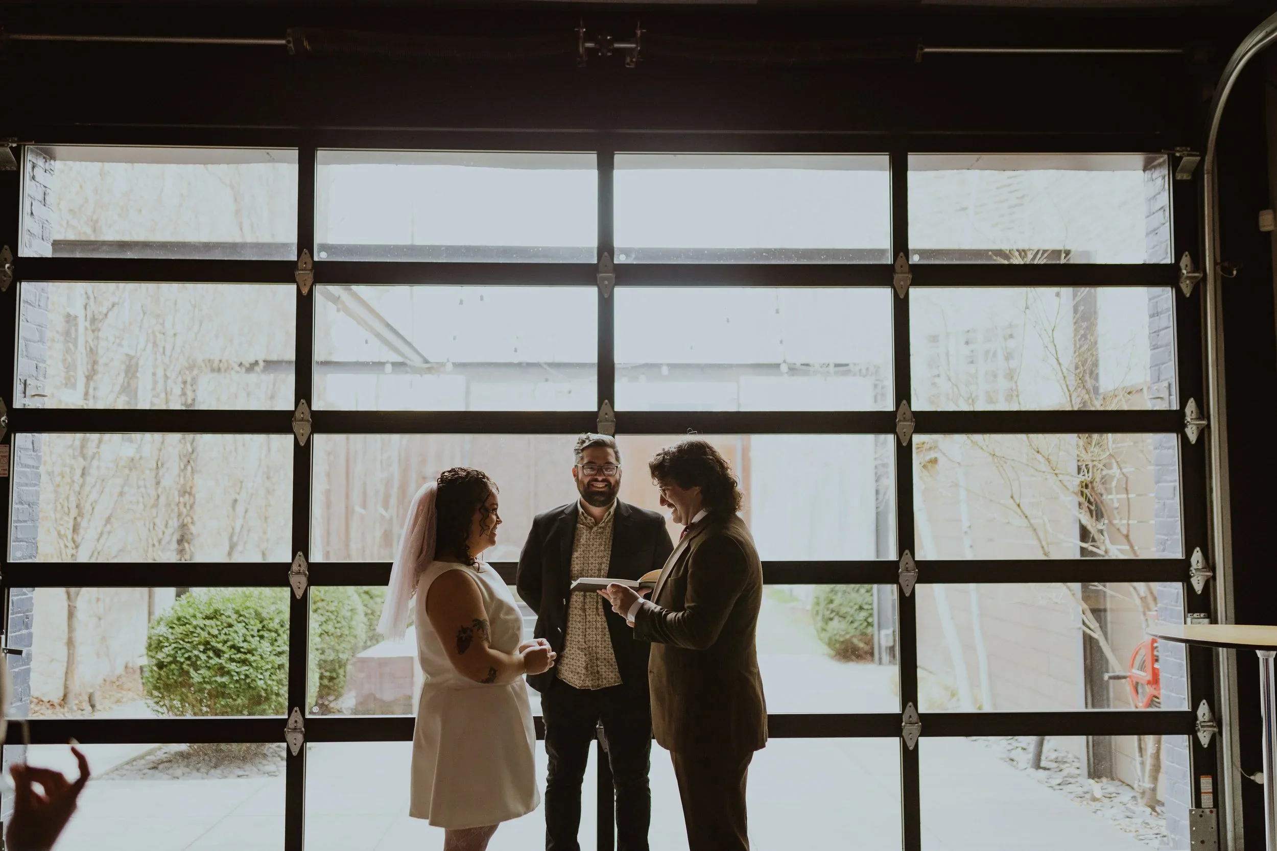 A wedding ceremony taking place inside a garage at the Lytle House in Chicago with large glass panel doors, with a woman in a white dress and veil, a man in a black suit, and another man in a brown blazer. Micro Wedding Images by Ttolbphotog