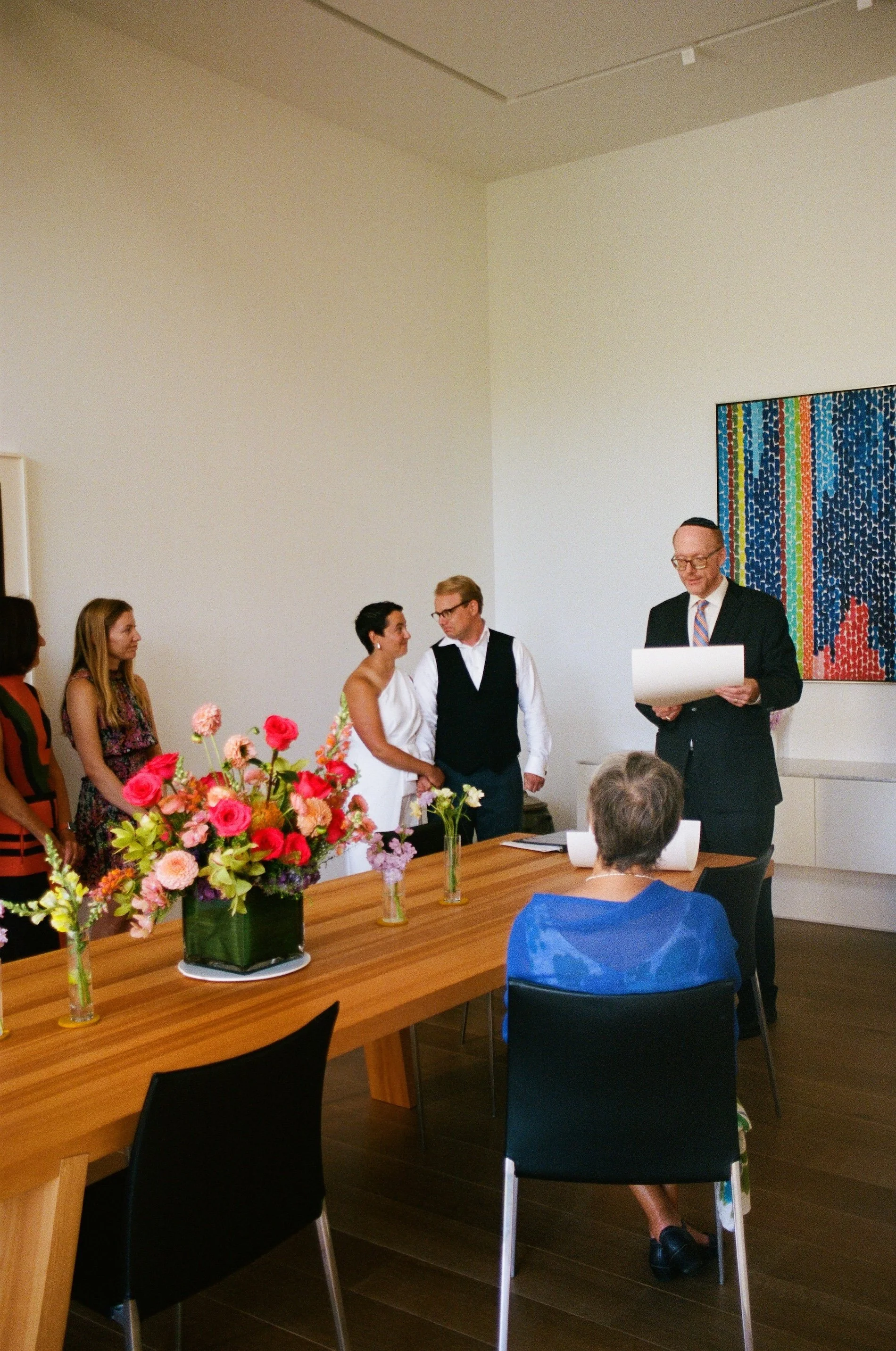 Micro Backyard Intimate Jewish Wedding in Chicago. A group of people standing in a room during a formal event, with a man in a suit reading from a paper, and a colorful abstract painting on the wall. Flower arrangements on a wooden table.