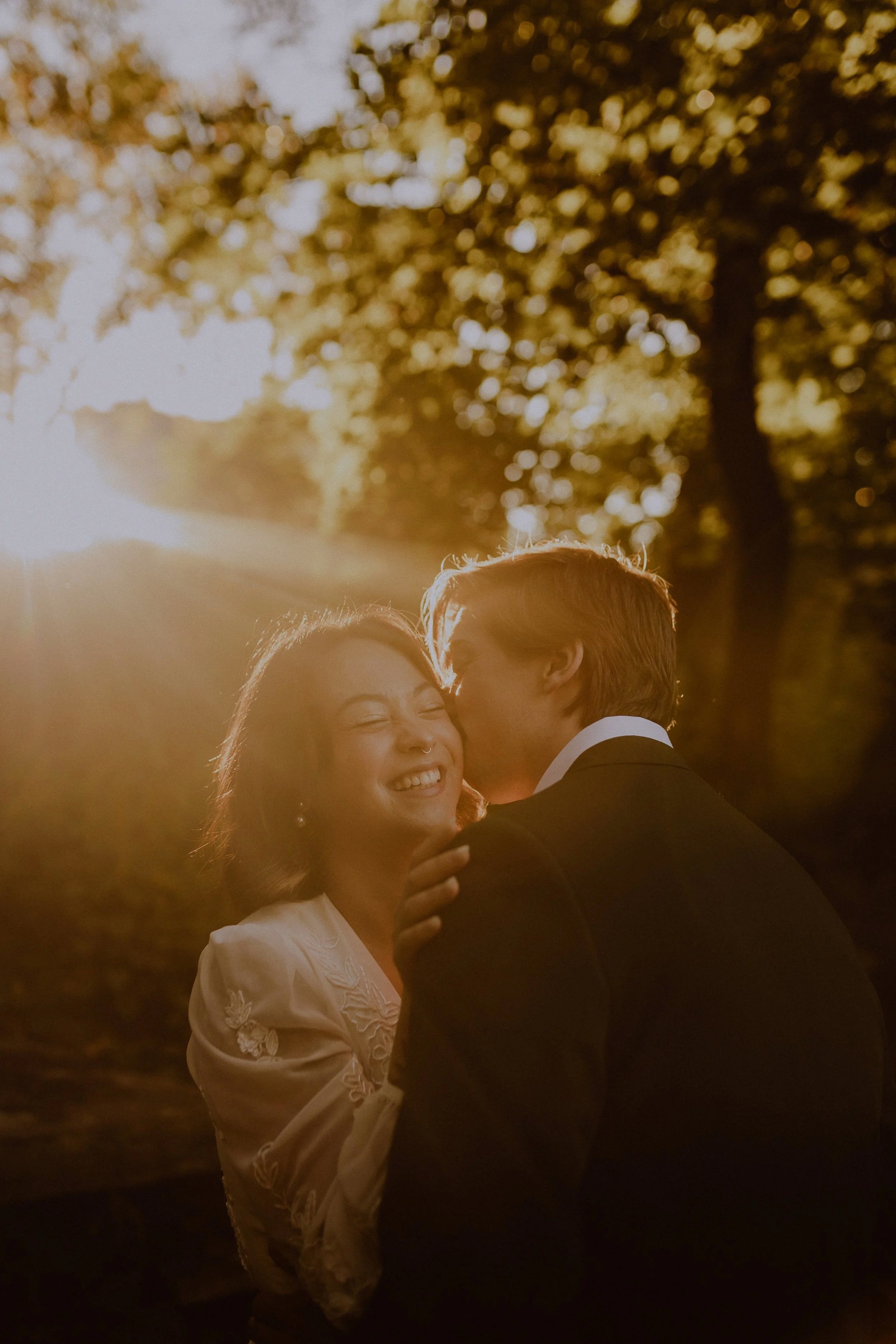 A couple sharing a kiss outdoors during sunset, with warm golden sunlight streaming through trees, the woman smiling happily. Lincoln Park Elopement at the Alfred Caldwell Lily Pool by Ttolbphotog